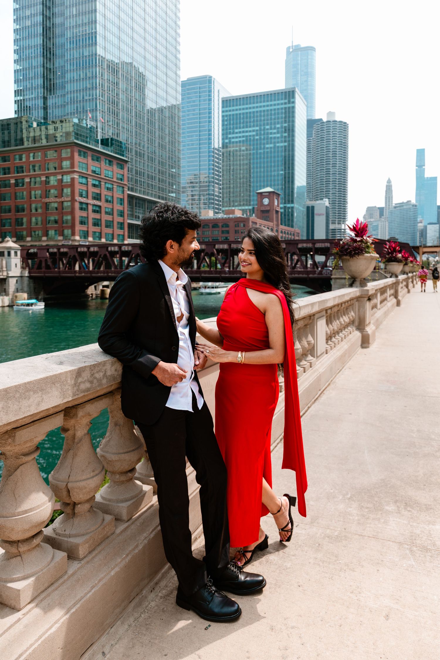 Couple in red dress and black suit posing on Chicago Riverwalk bridge with city skyline backdrop.