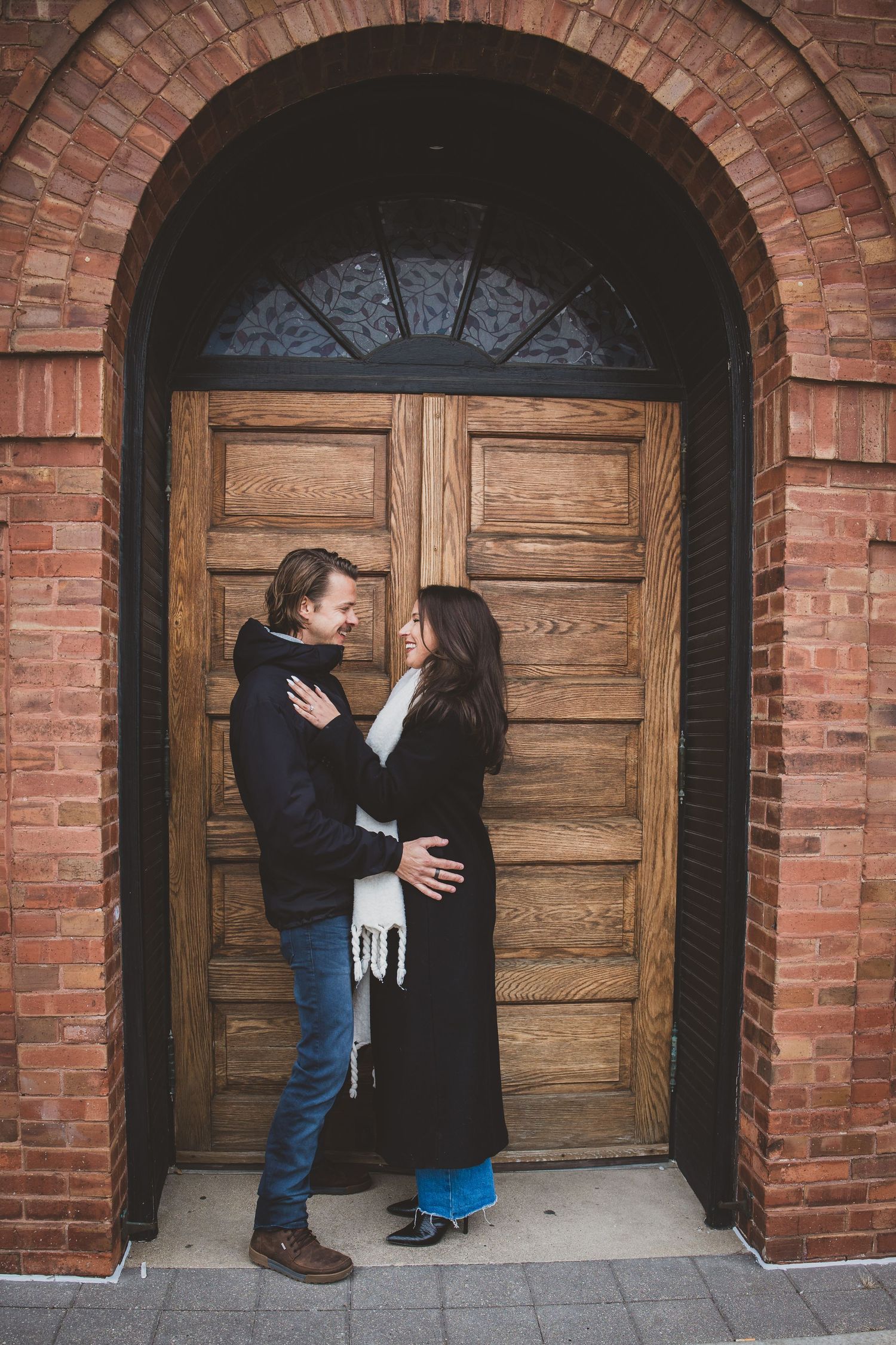 Couple embracing and smiling at each other in front of a wooden arched door set in a red brick wall.