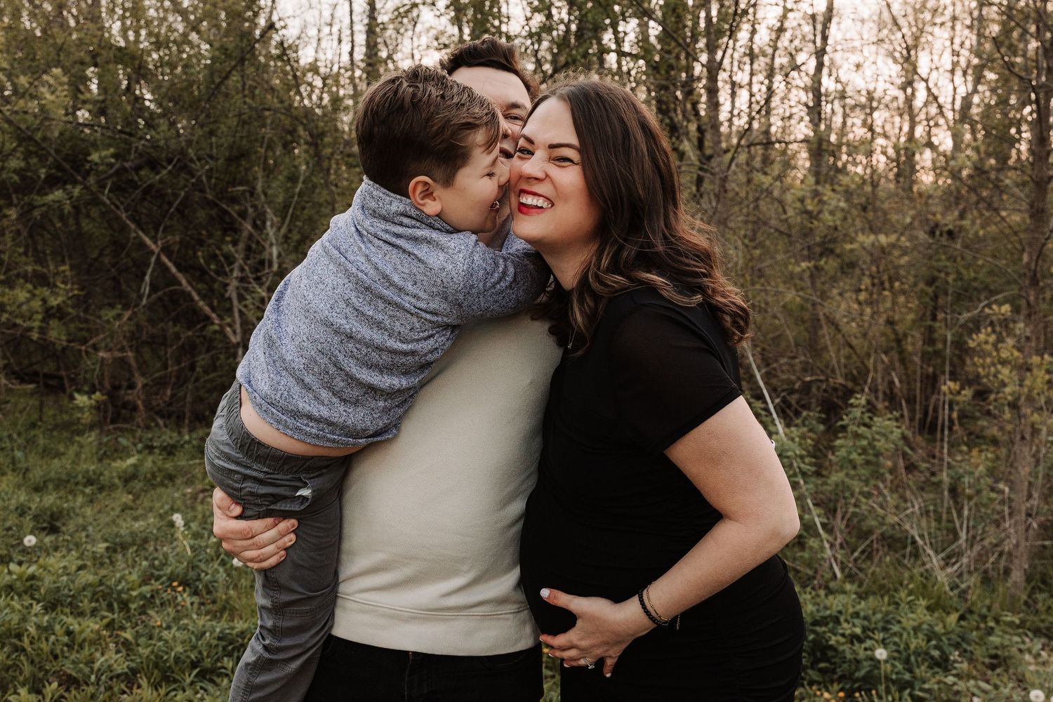 Happy family maternity photo with child kissing mom outdoors in green nature setting.