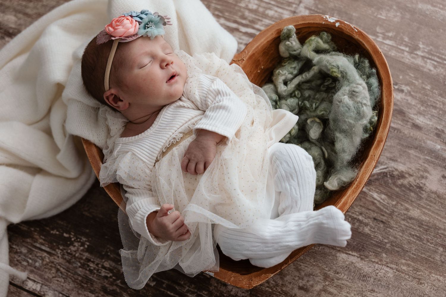 Newborn baby girl in white knit outfit with floral headband sleeping in a heart-shaped wooden bowl.