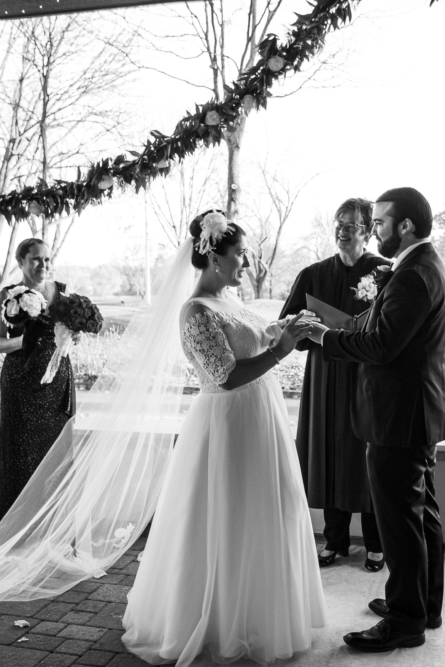 Black and white photo of bride and groom exchanging vows under floral arch at outdoor ceremony.