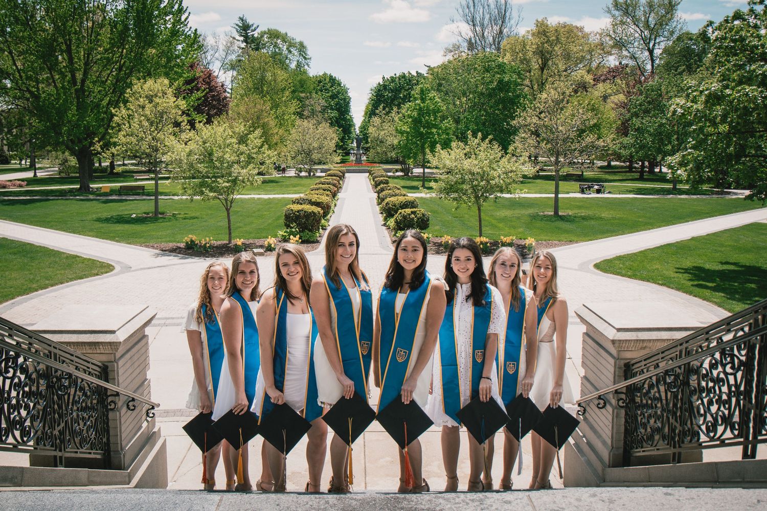 Group of female graduates in white dresses and teal sashes posing on ornate steps in a lush green university campus.