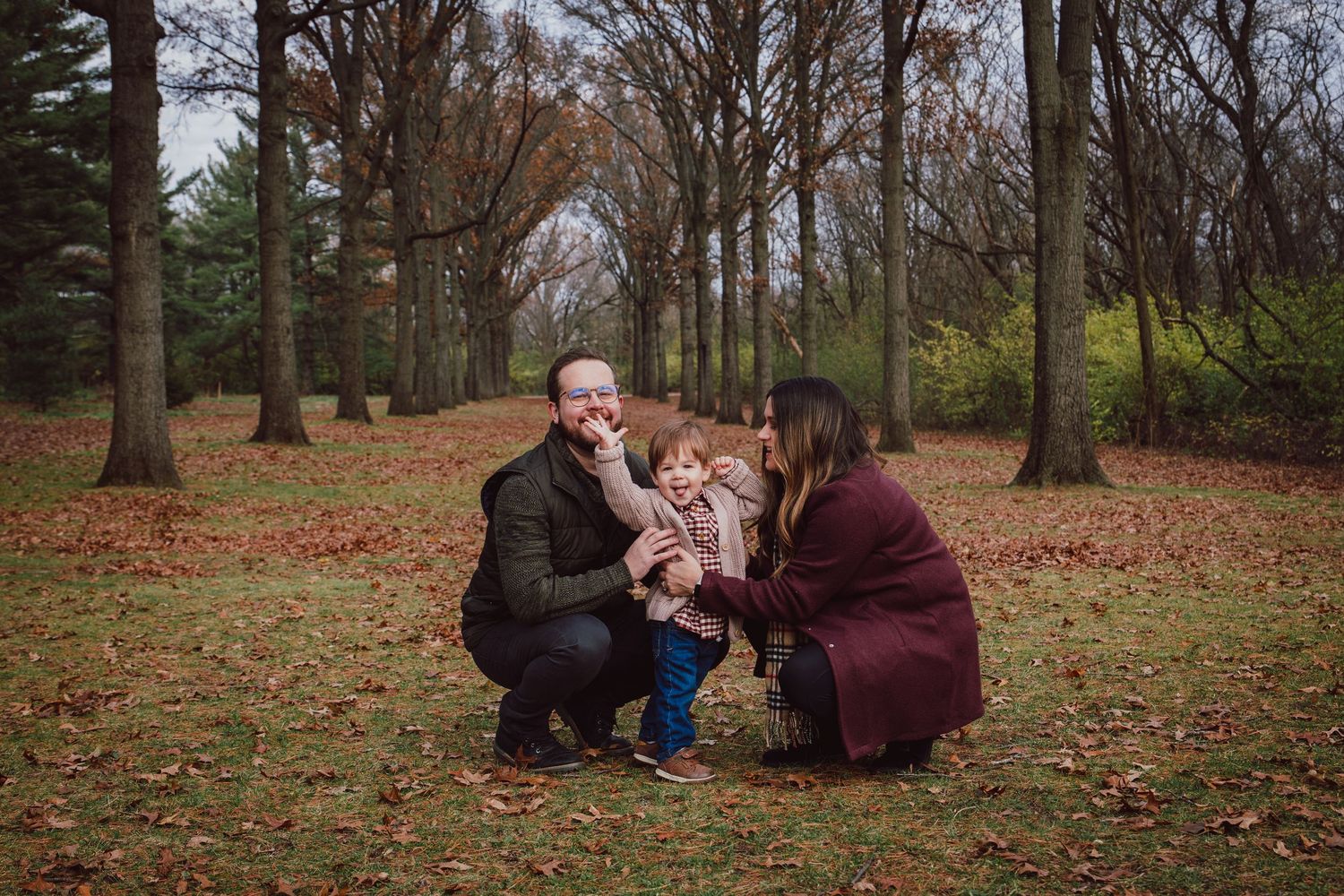 Parents crouching with toddler in autumn park covered in fallen leaves.