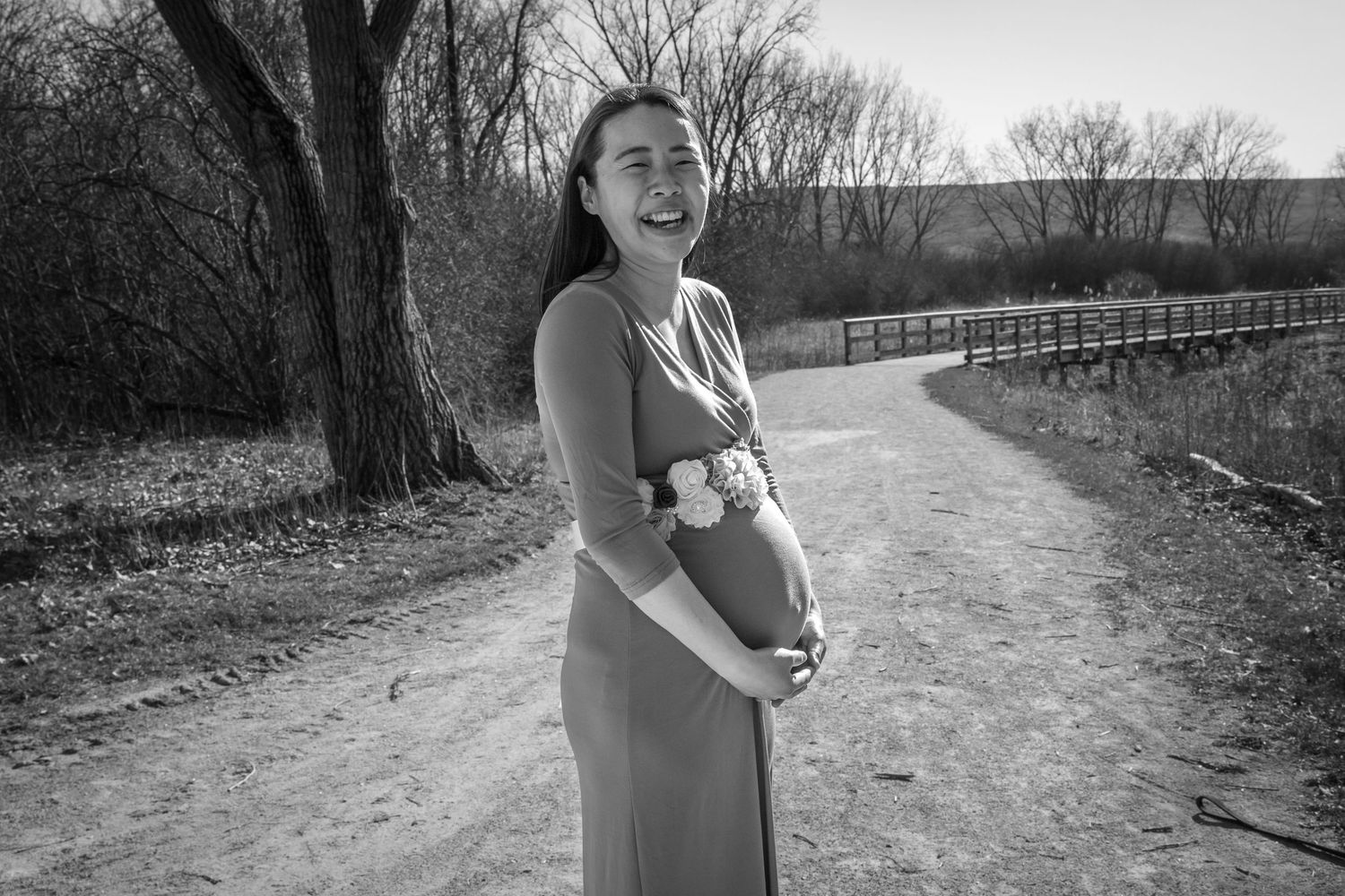 Black and white photo of a smiling pregnant woman in a dress holding her belly on a dirt path.
