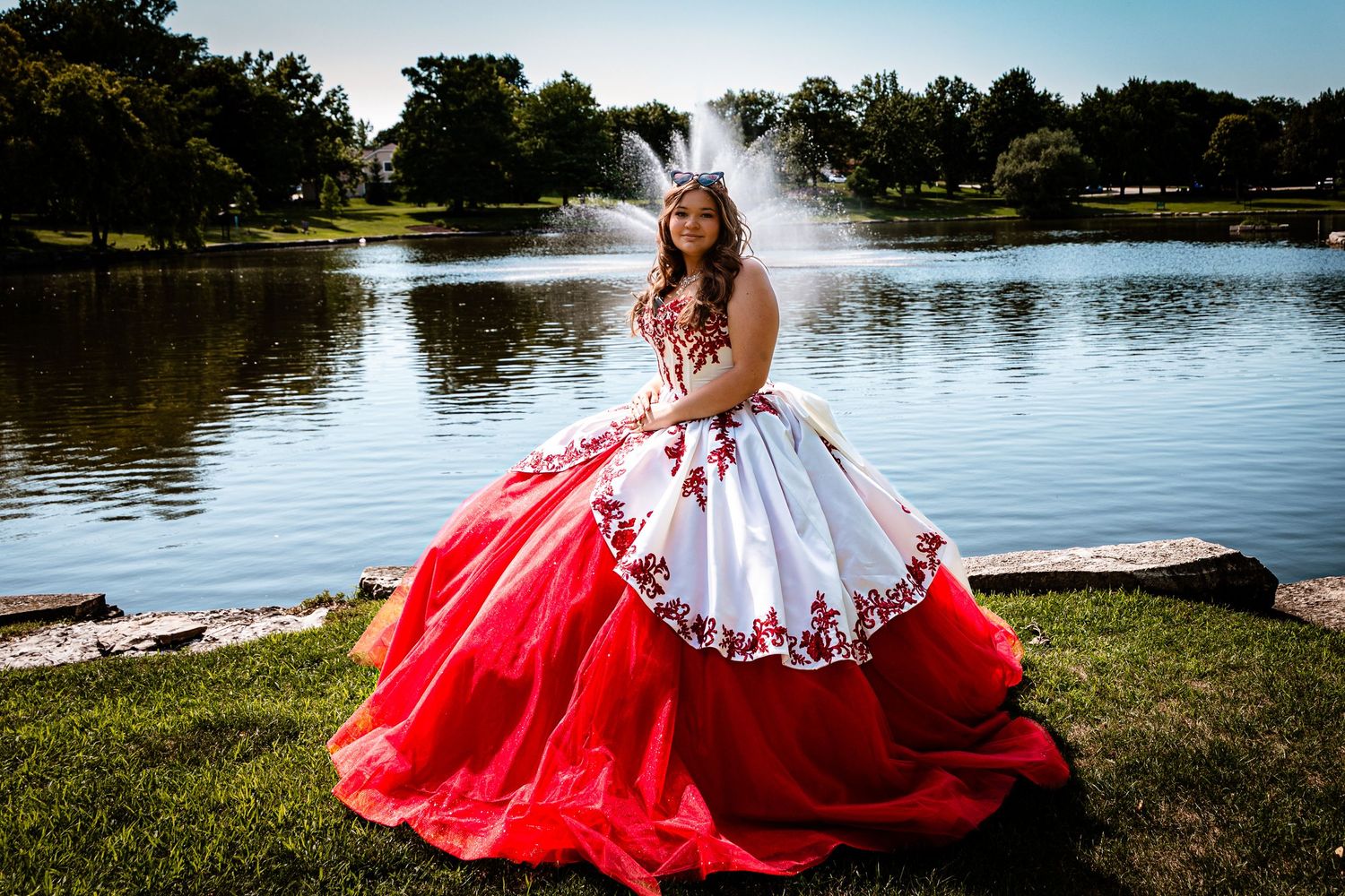 Young woman in red and white quinceañera ball gown poses by a lake with a fountain in the background.