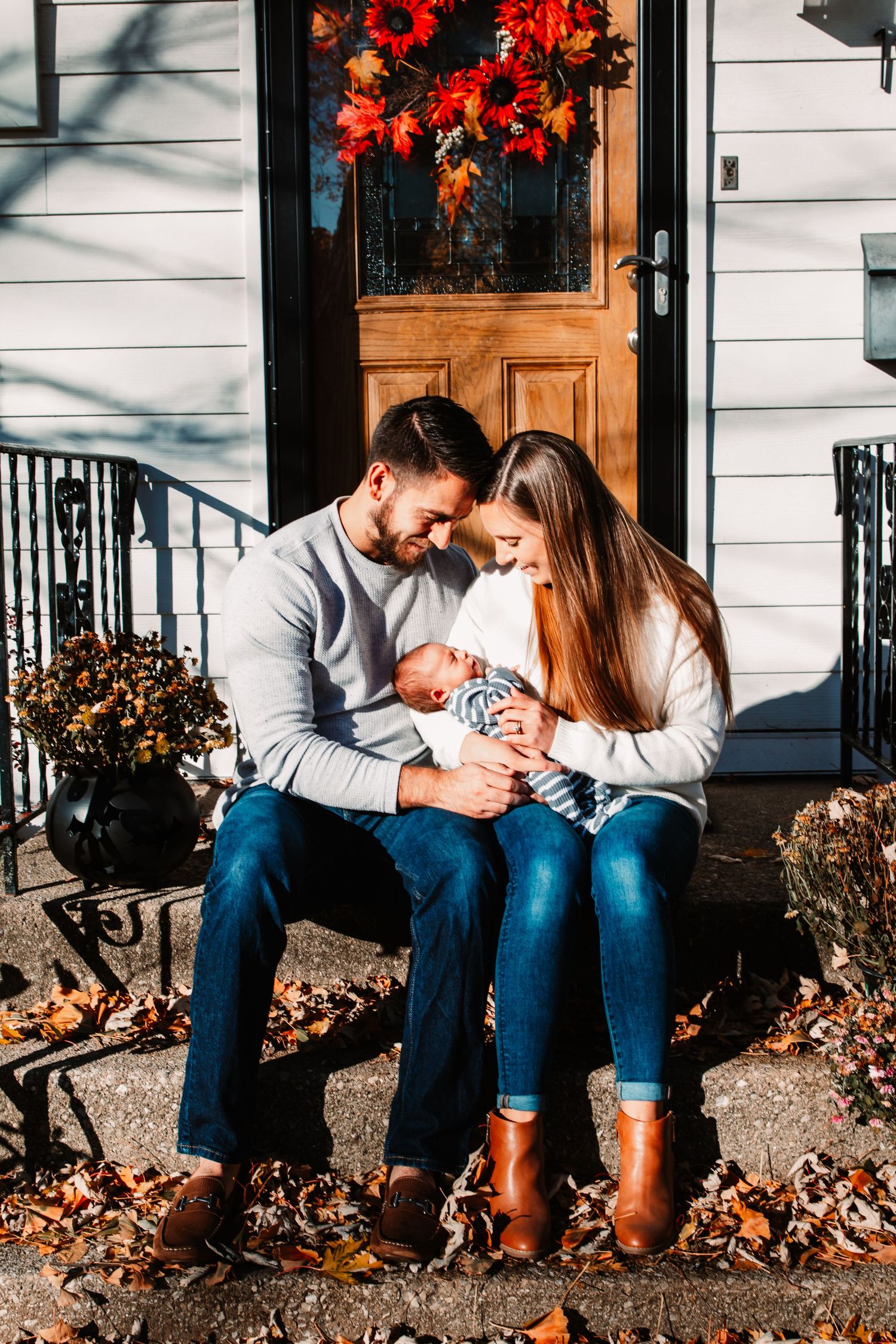 Young couple sitting on front porch steps in autumn, lovingly cradling their newborn baby.