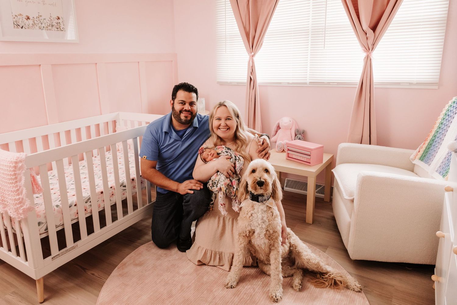 Family with newborn baby and golden doodle dog posing in a pink nursery room.