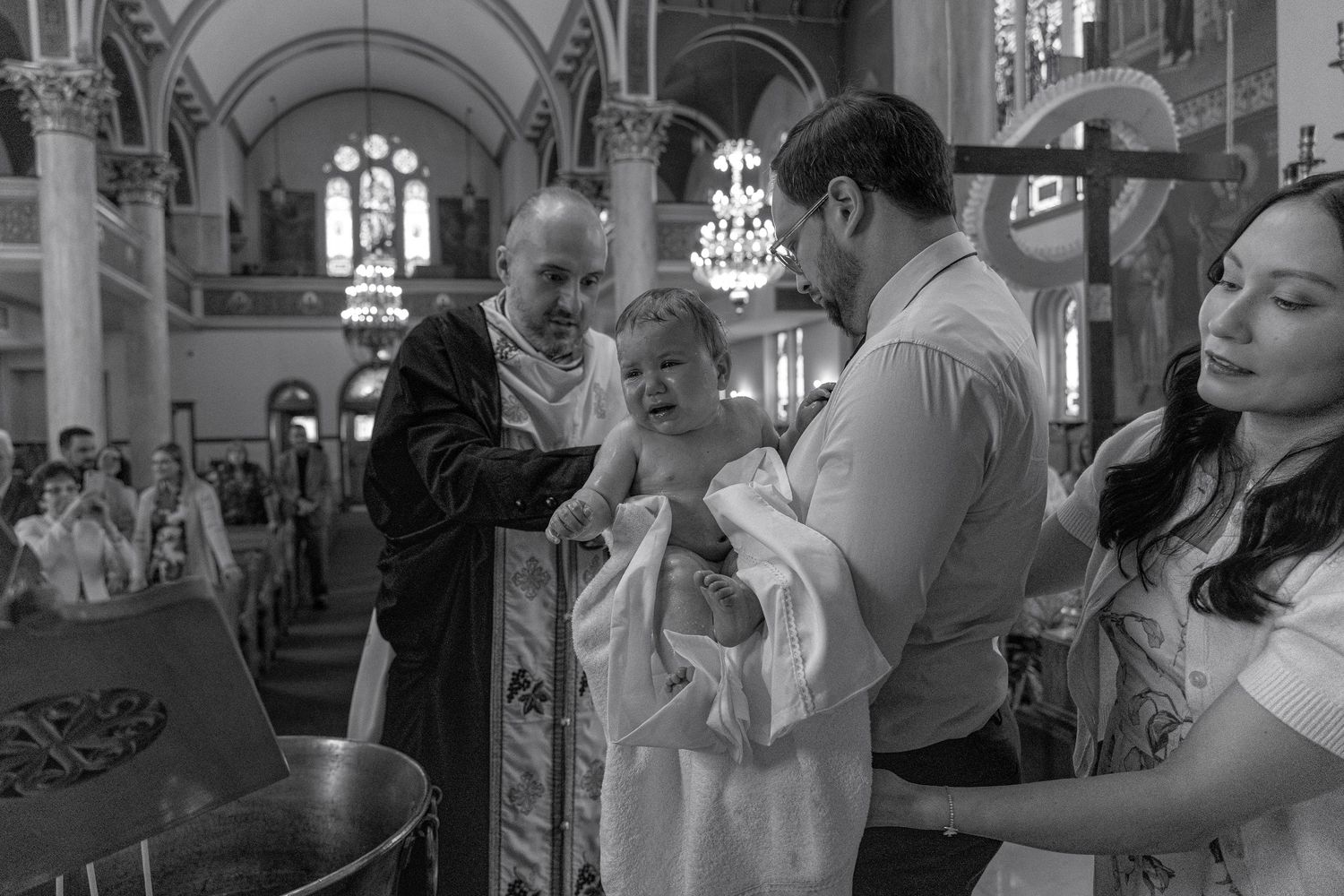 Black and white photo of Orthodox priest baptizing an infant held by parents in a grand church.
