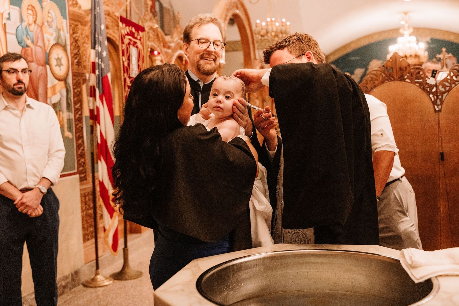 Orthodox baptism ceremony with priest anointing baby over baptismal font surrounded by family members.