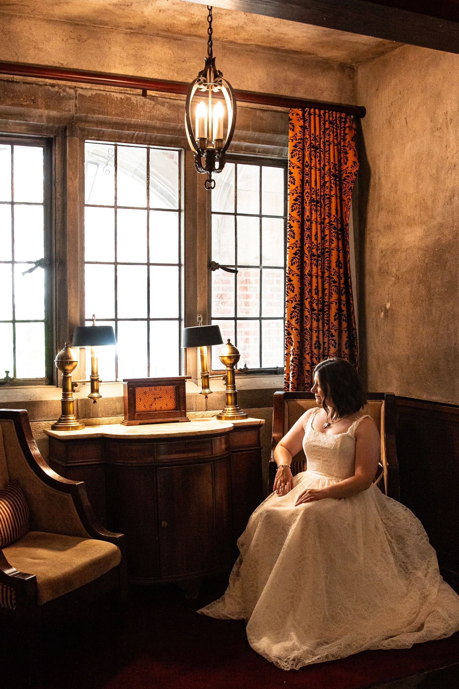 Bride in white gown sits pensively by vintage desk with brass lamps in a warmly lit rustic room.