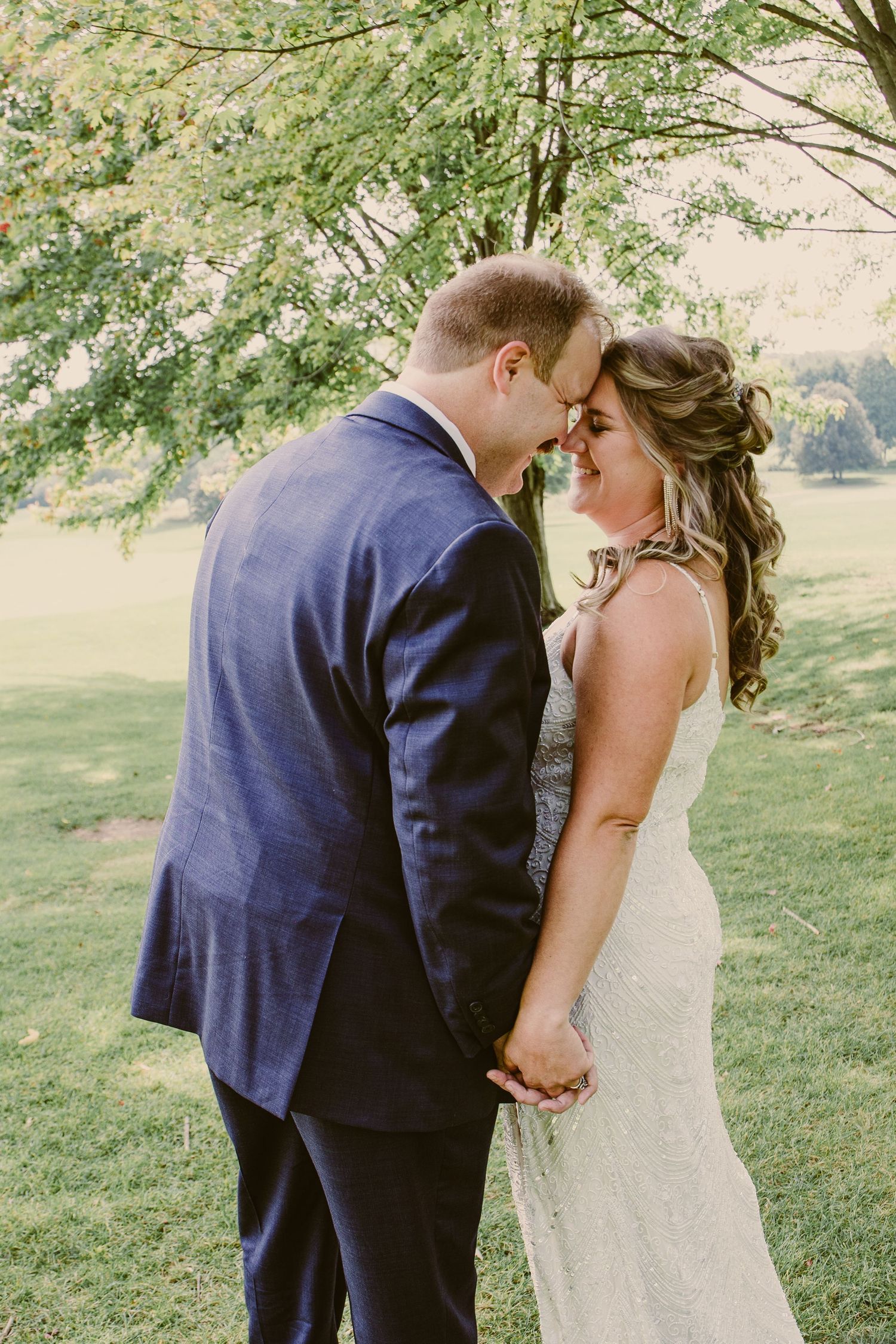 A bride and groom touch foreheads tenderly while holding hands in a lush green outdoor setting.