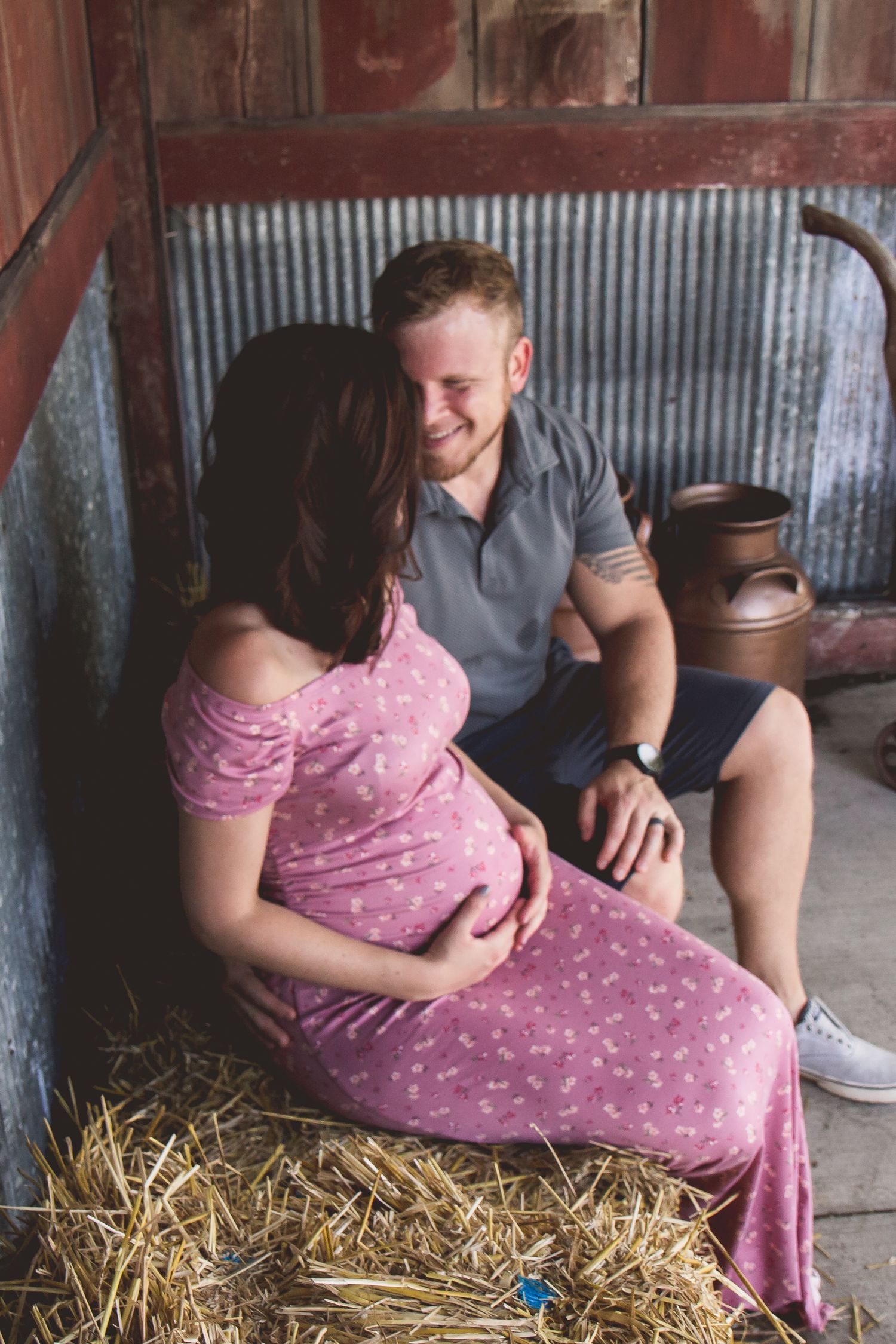 A smiling couple sitting on hay in a rustic barn, with the woman in a pink maternity dress.