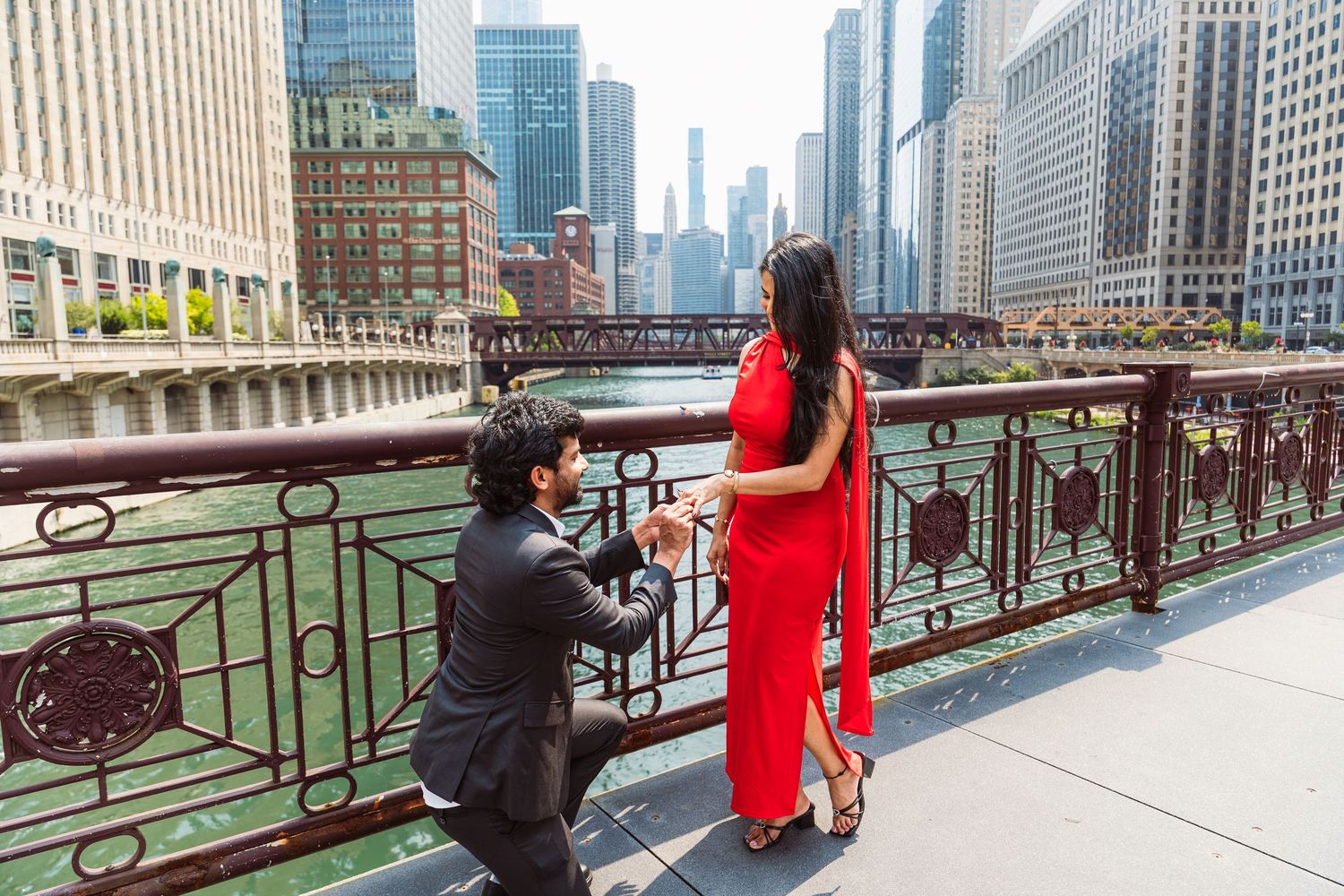 Man proposing to woman in red dress on Chicago Riverwalk bridge with city skyline backdrop.