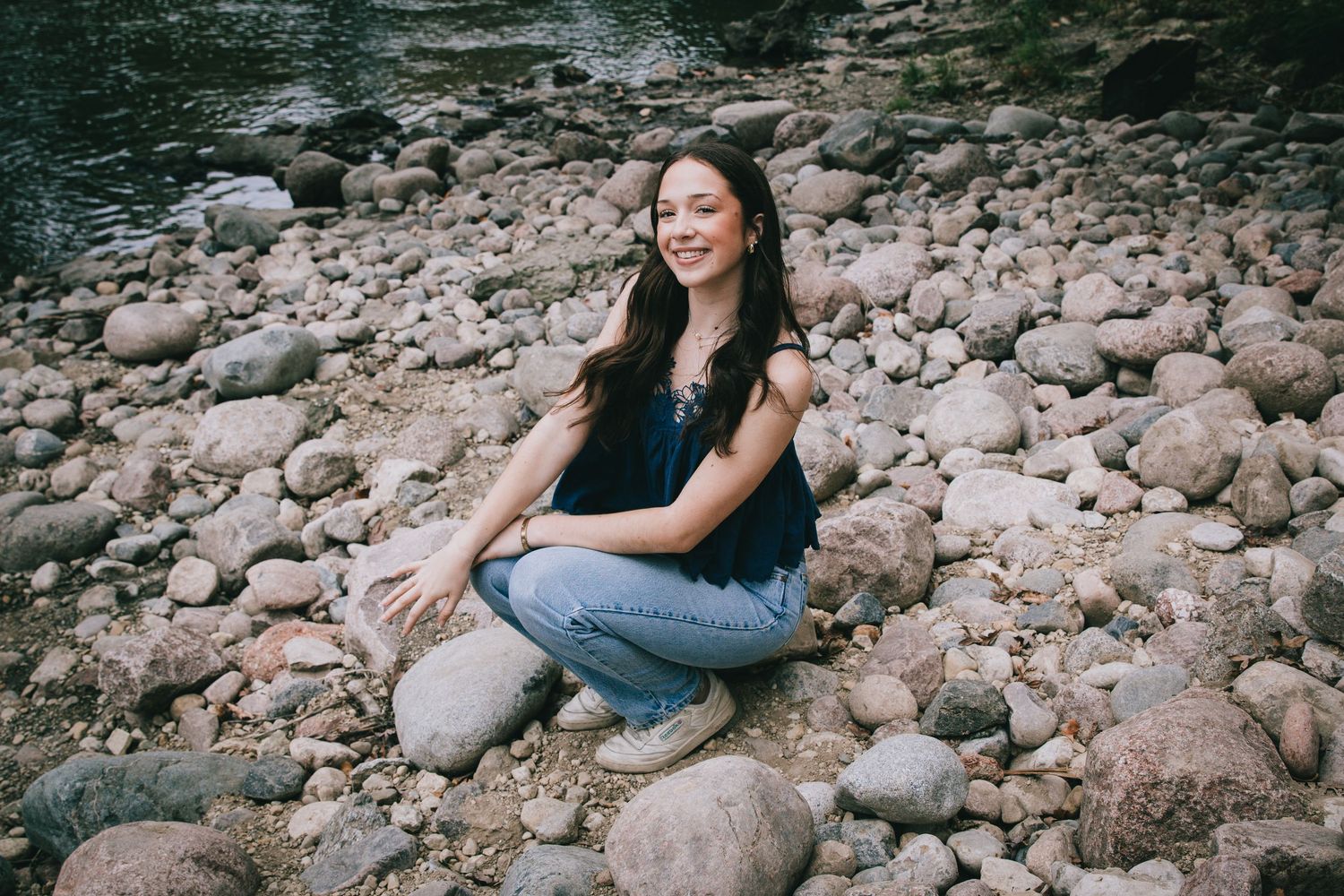 Young woman with long dark hair smiling while sitting on rocky riverbank in casual jeans and navy top.