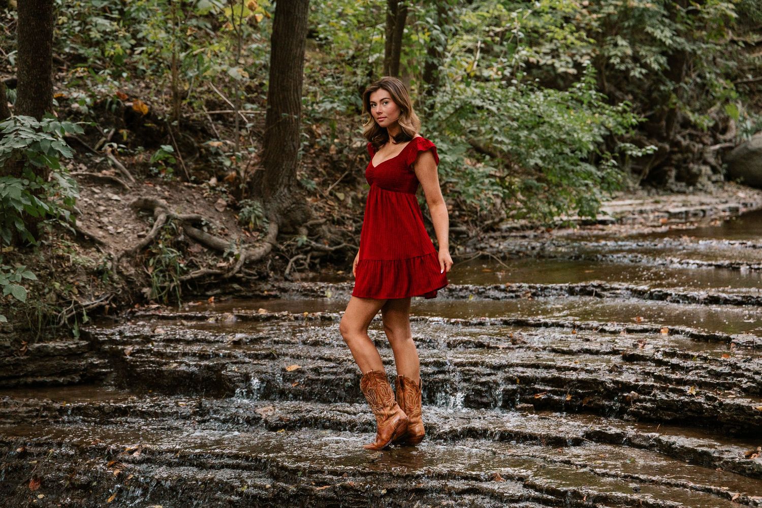 Woman in red dress and brown cowboy boots standing in a shallow forest creek surrounded by lush green trees.
