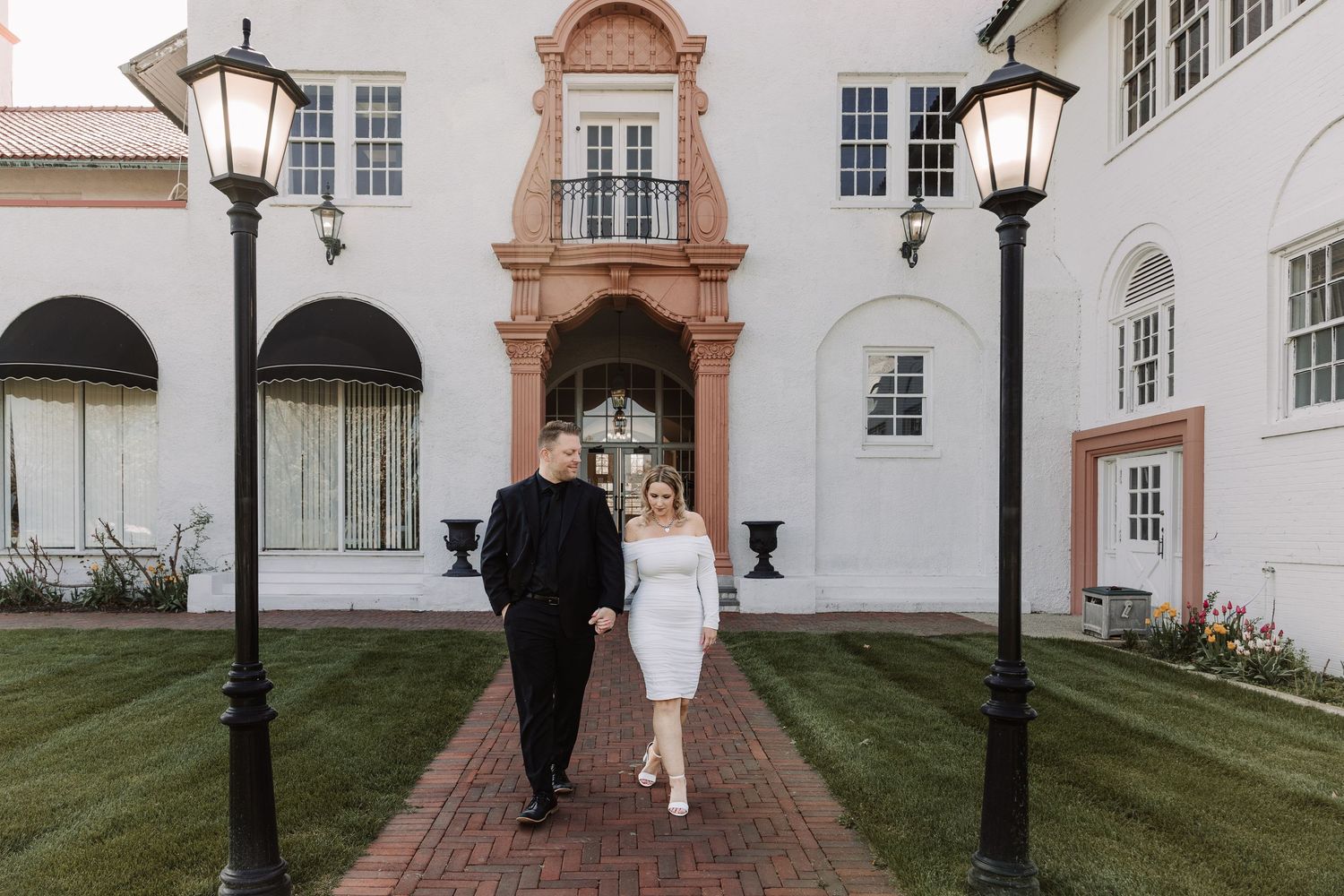 Couple holding hands walking toward camera outside elegant white building with brick pathway and ornate lamp posts.