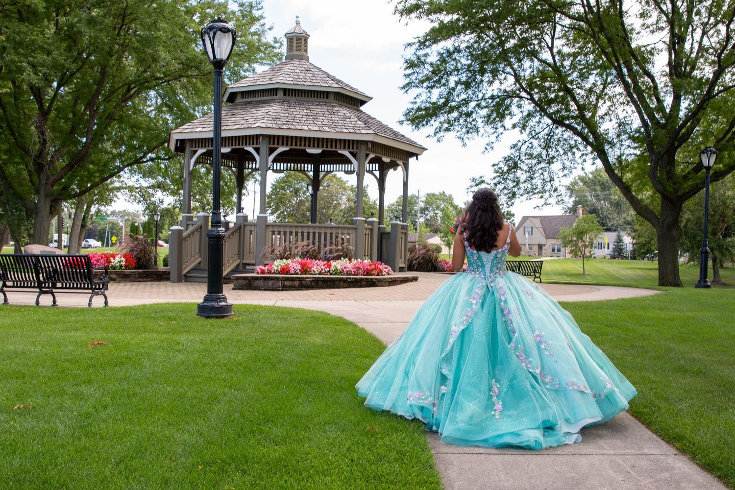Girl in teal quinceañera ball gown walks along park path toward a classic wooden gazebo surrounded by lush green trees.