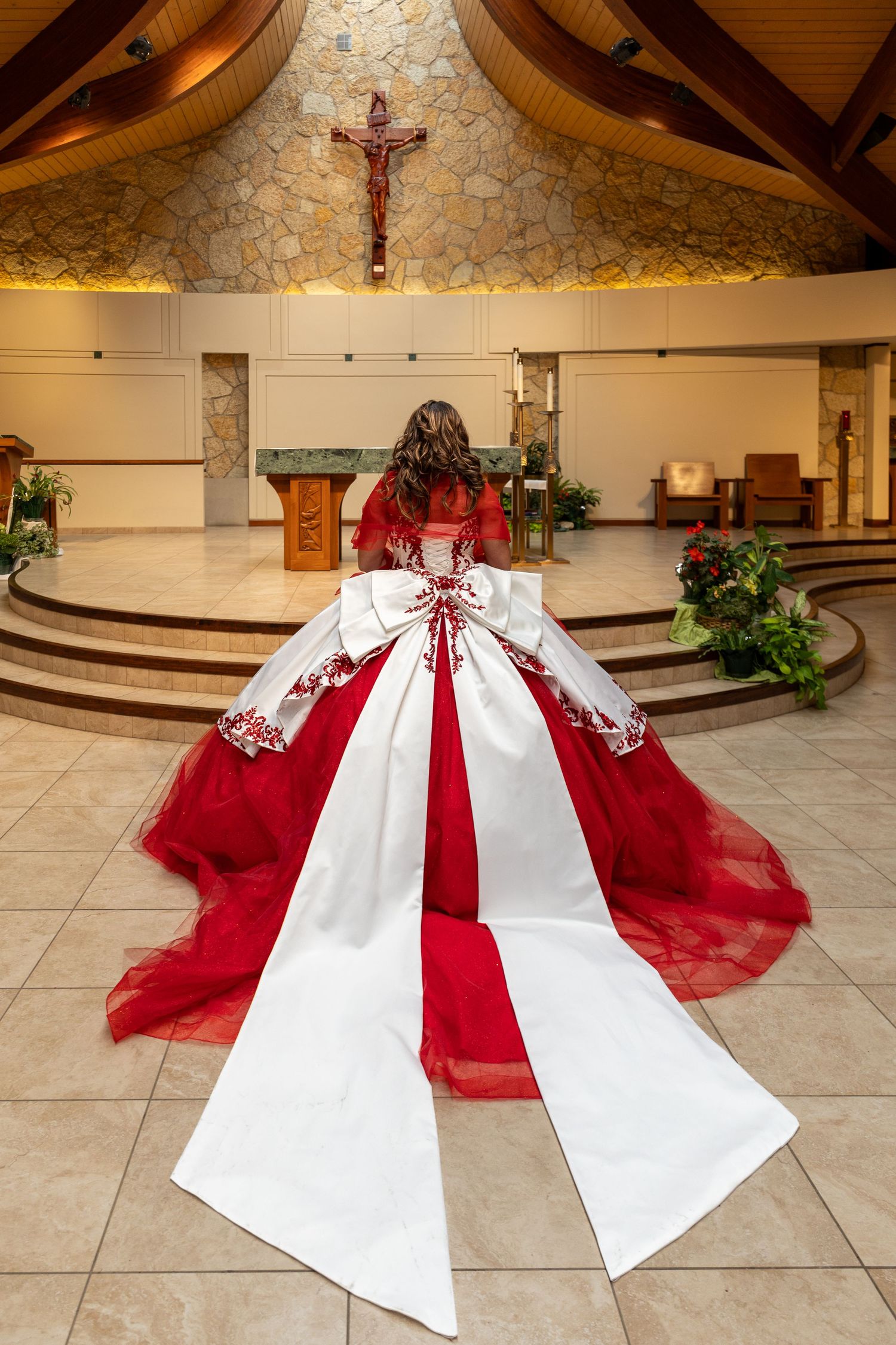 Quinceañera girl in white and red ball gown with large bow kneels before church altar beneath a crucifix.