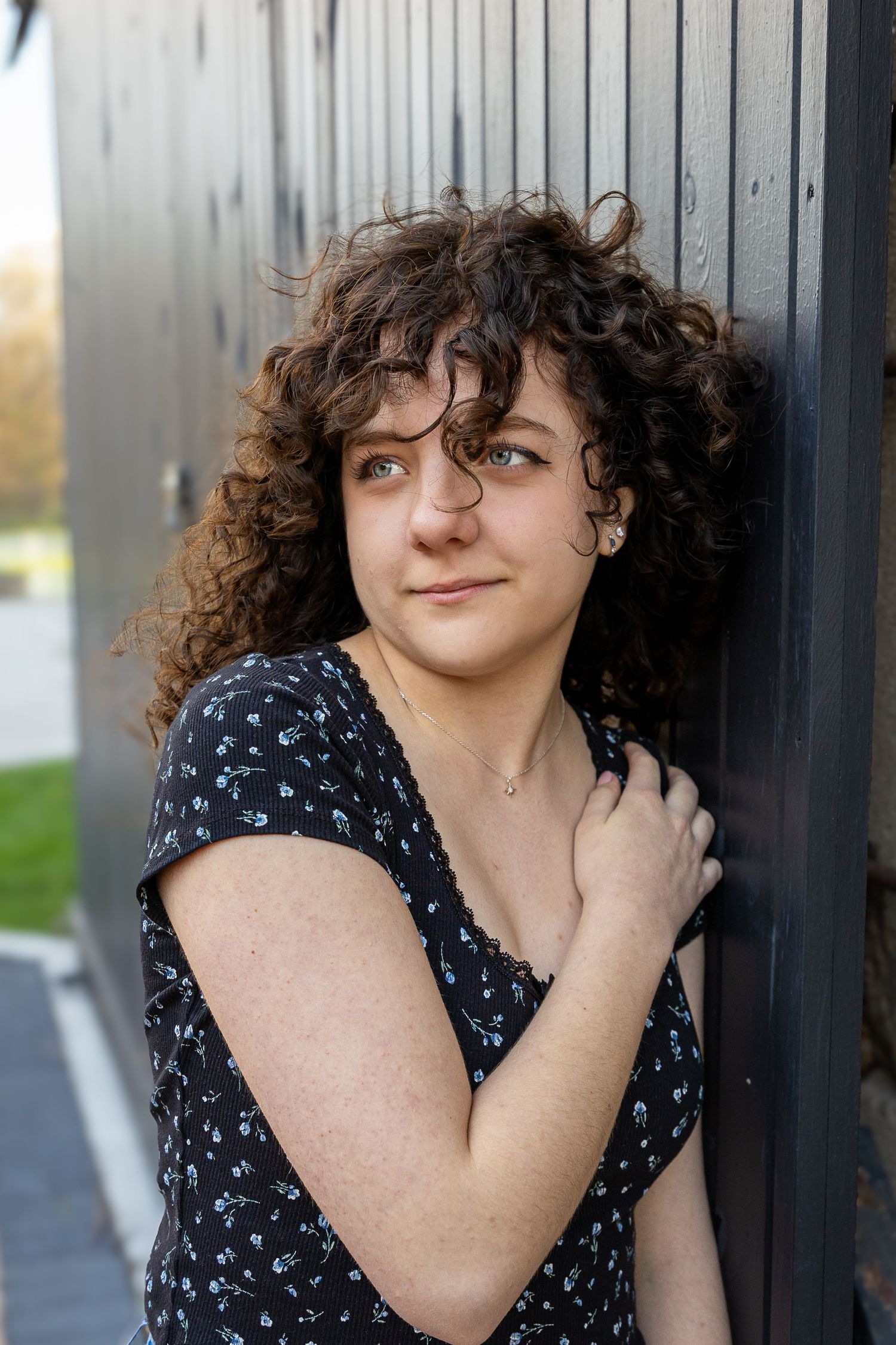 Young woman with curly brown hair leaning against a dark wall, wearing a floral dress, smiling softly outdoors.