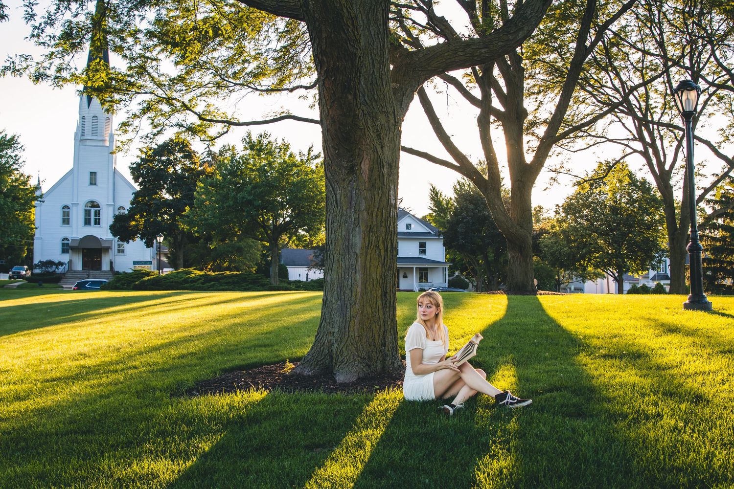 Woman reading under a large tree on a sunny green lawn beside a white church in a peaceful park setting.
