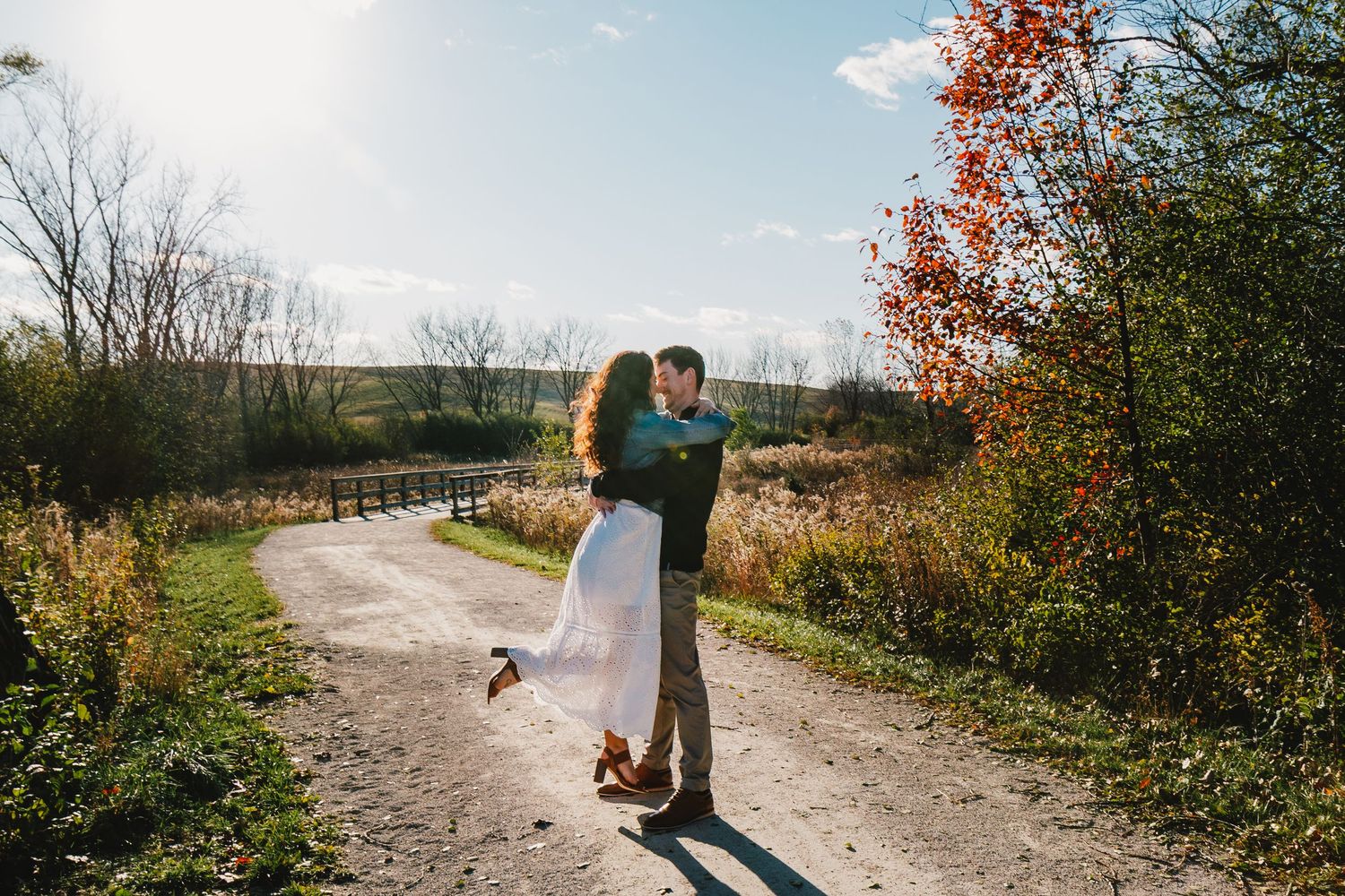 Couple embracing on a sunny autumn dirt road surrounded by colorful fall foliage and open fields.