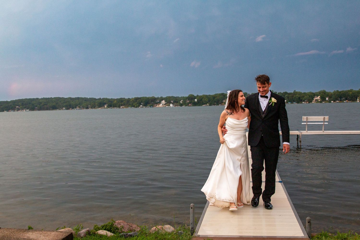 A bride and groom walk hand in hand along a dock with a serene lake and dramatic sky behind them.