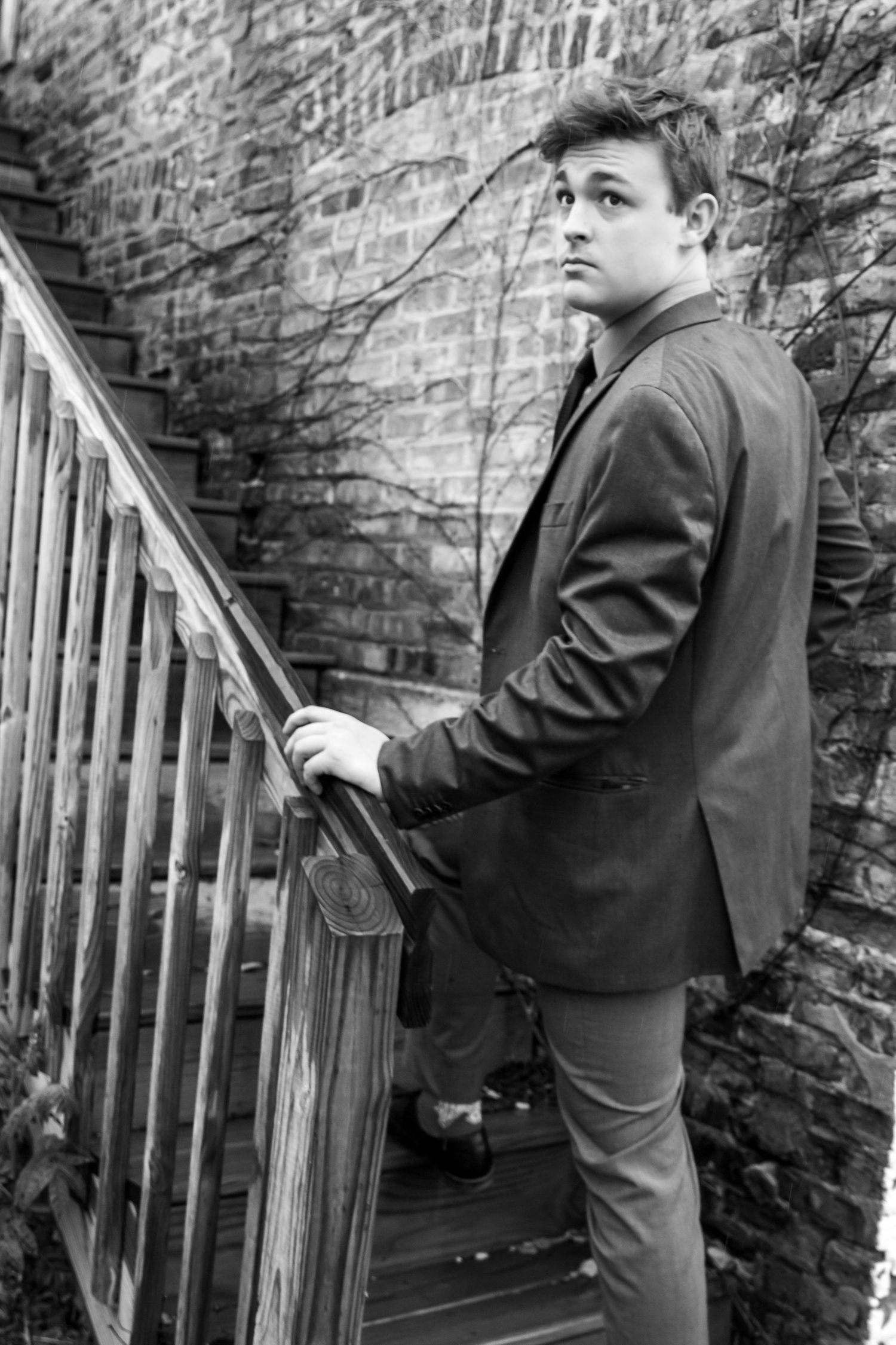 Black and white photo of a young man in a suit standing on wooden stairs against a brick wall.