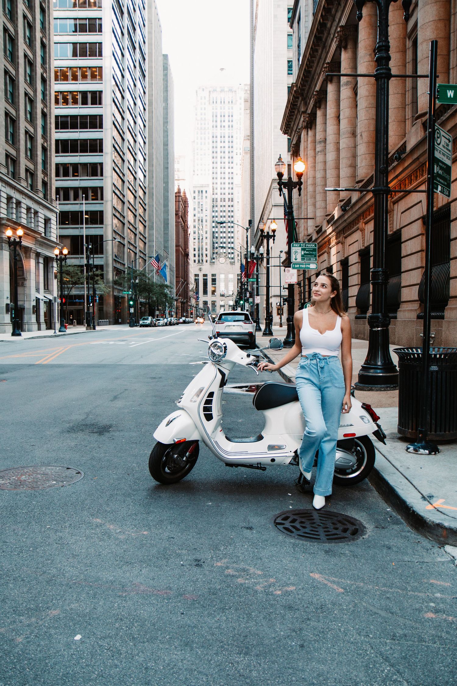 Young woman in white top and jeans leaning on a white Vespa scooter on a quiet urban street lined with tall buildings.