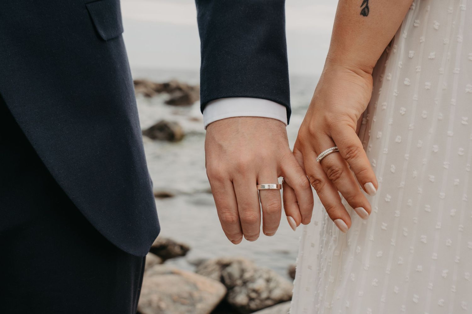 Close-up of newlywed couple's hands showing wedding rings against a beach backdrop.