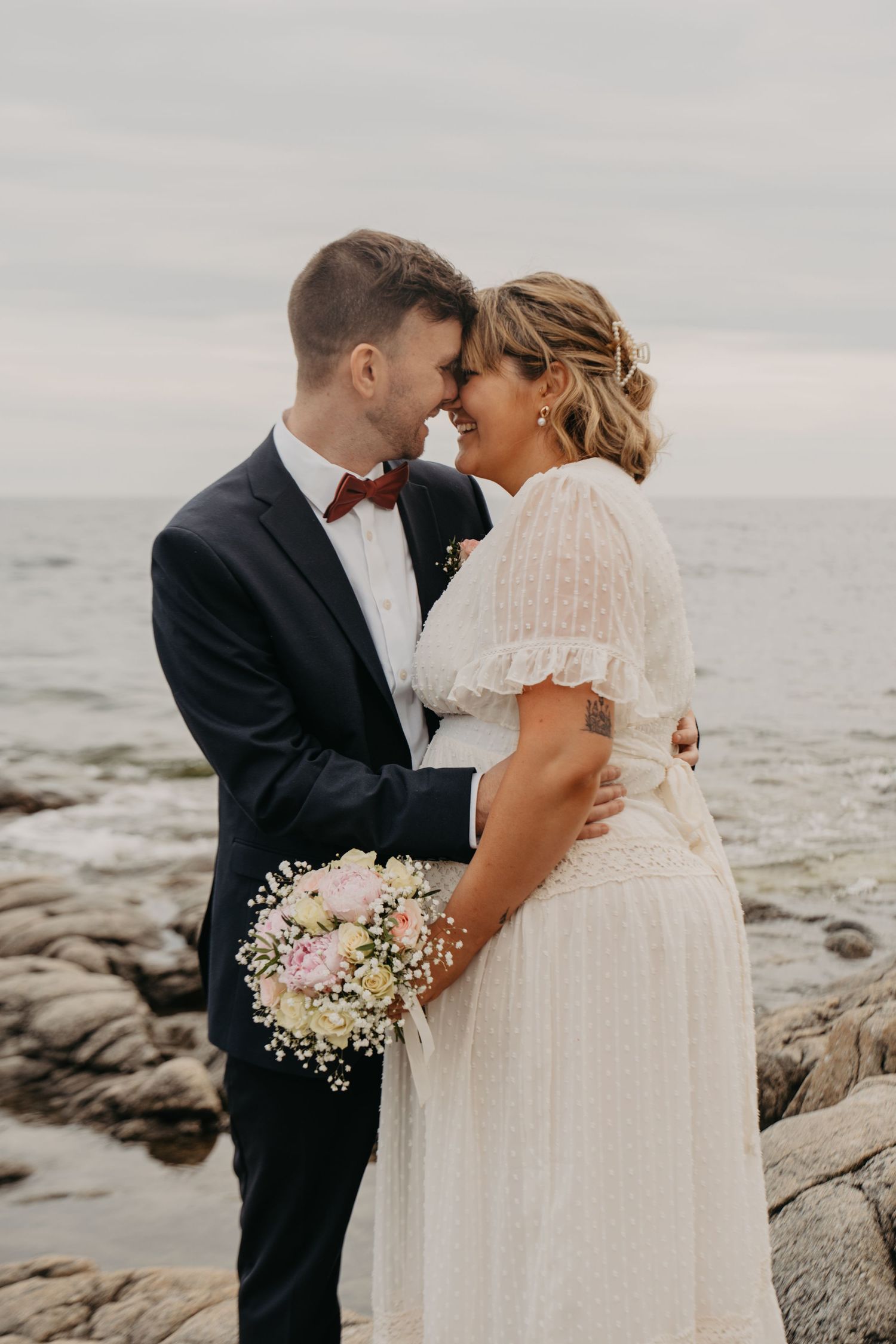 Romantic coastal wedding photos of couple embracing on rocky shoreline with bouquet of baby's breath.