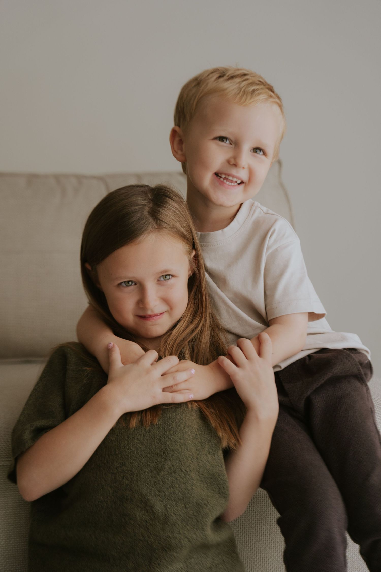 Two children in casual attire share playful moments on a couch against a neutral background.