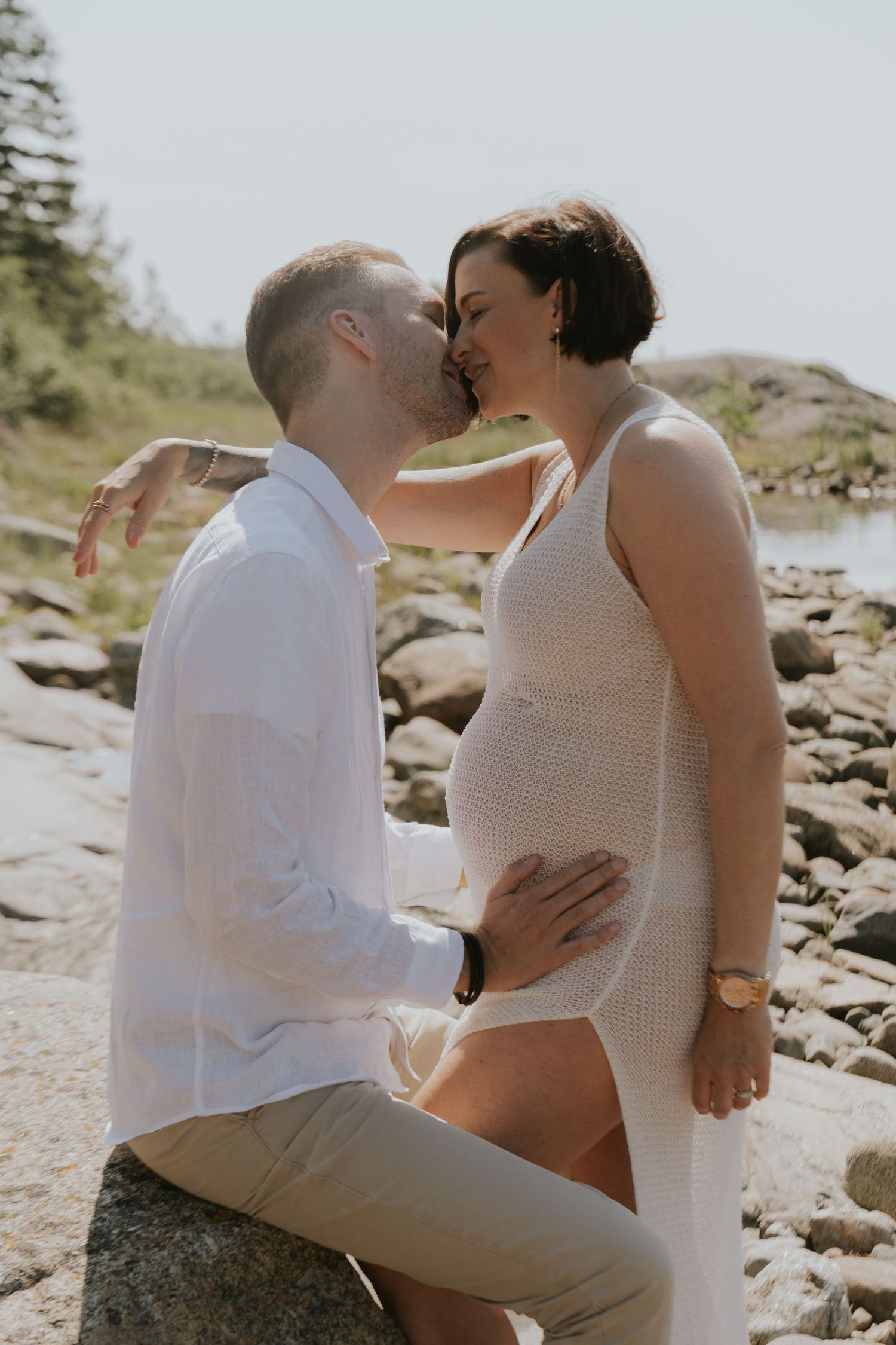 A couple shares tender moments while sitting on rocky coastal terrain during a sunny day.