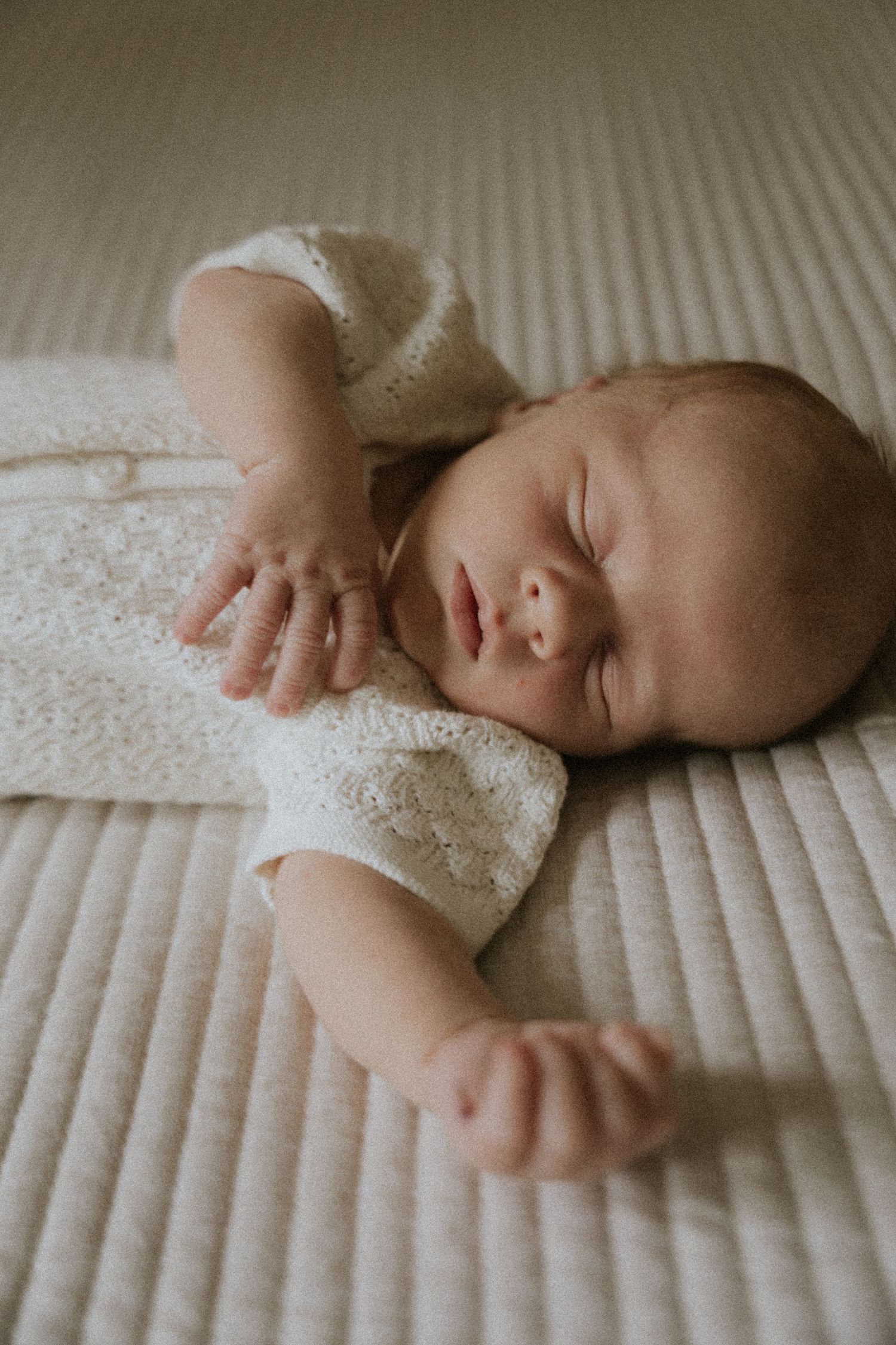 Sleeping newborn baby in white swaddle lying on textured cream bedding in soft natural light.
