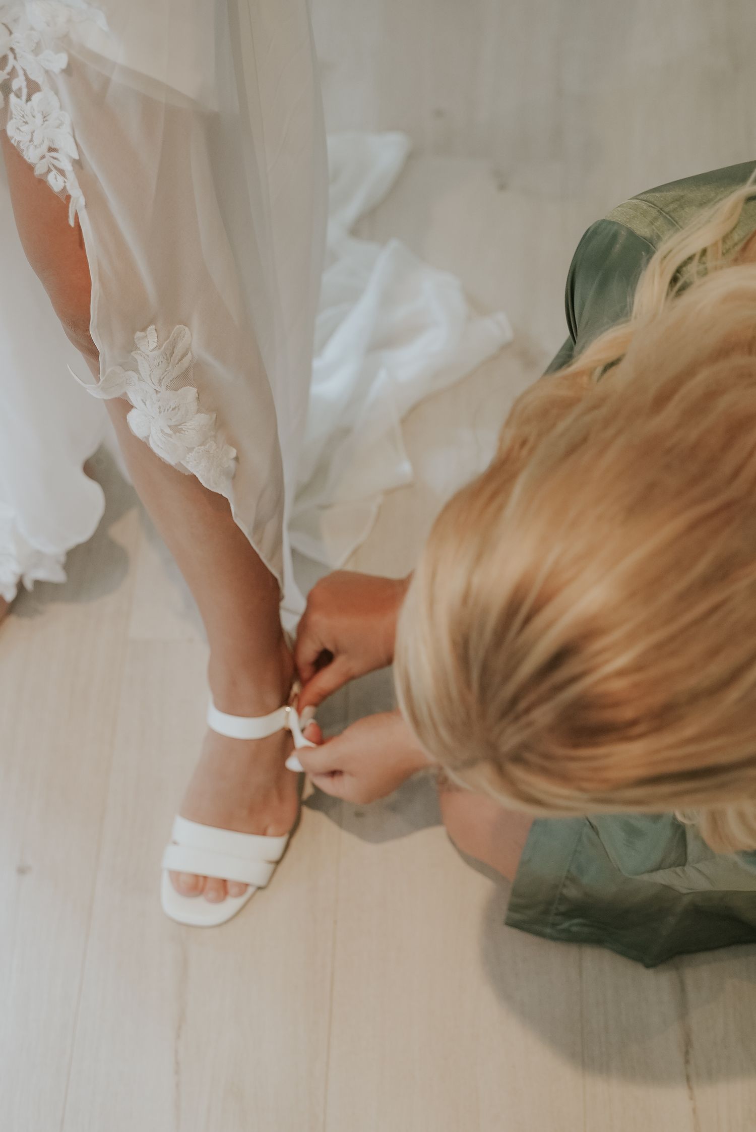 A close-up view of white bridal shoes being put on while wearing a sage green bridesmaid dress.