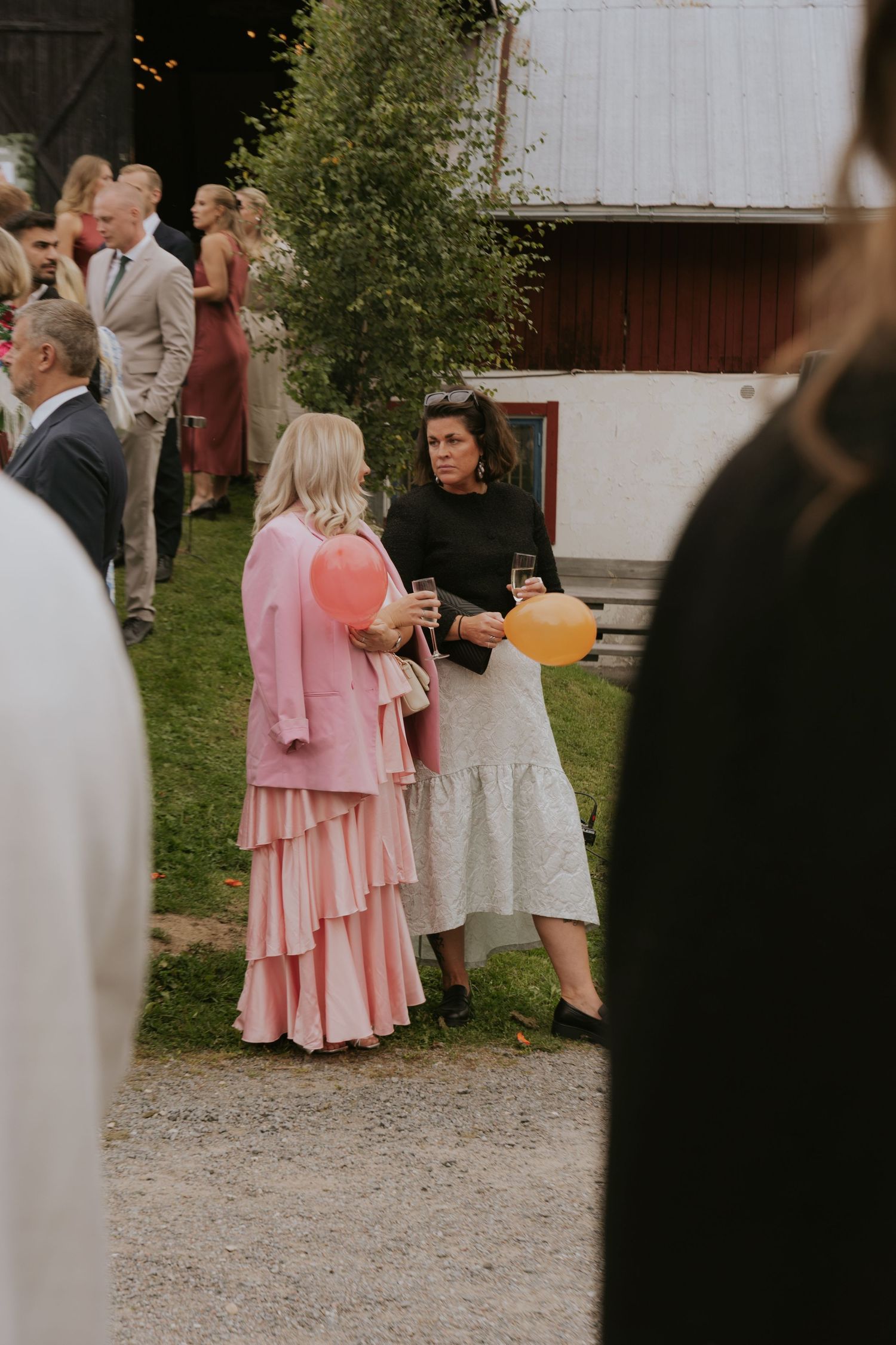 Wedding guests in pink and white dresses gather outside a rustic venue during an outdoor celebration.