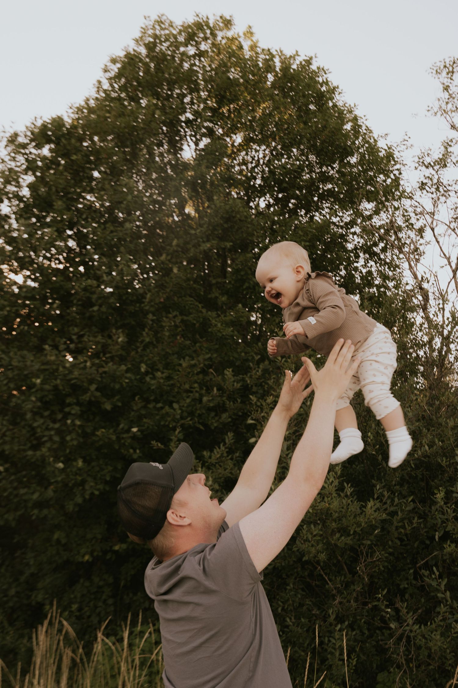 A playful moment captured as a baby is lifted high into the air against a backdrop of trees.