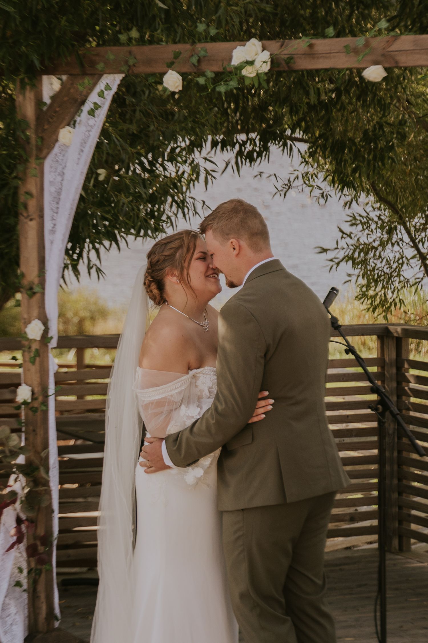 Romantic outdoor wedding ceremony under wooden pergola decorated with white fabric and flowers.