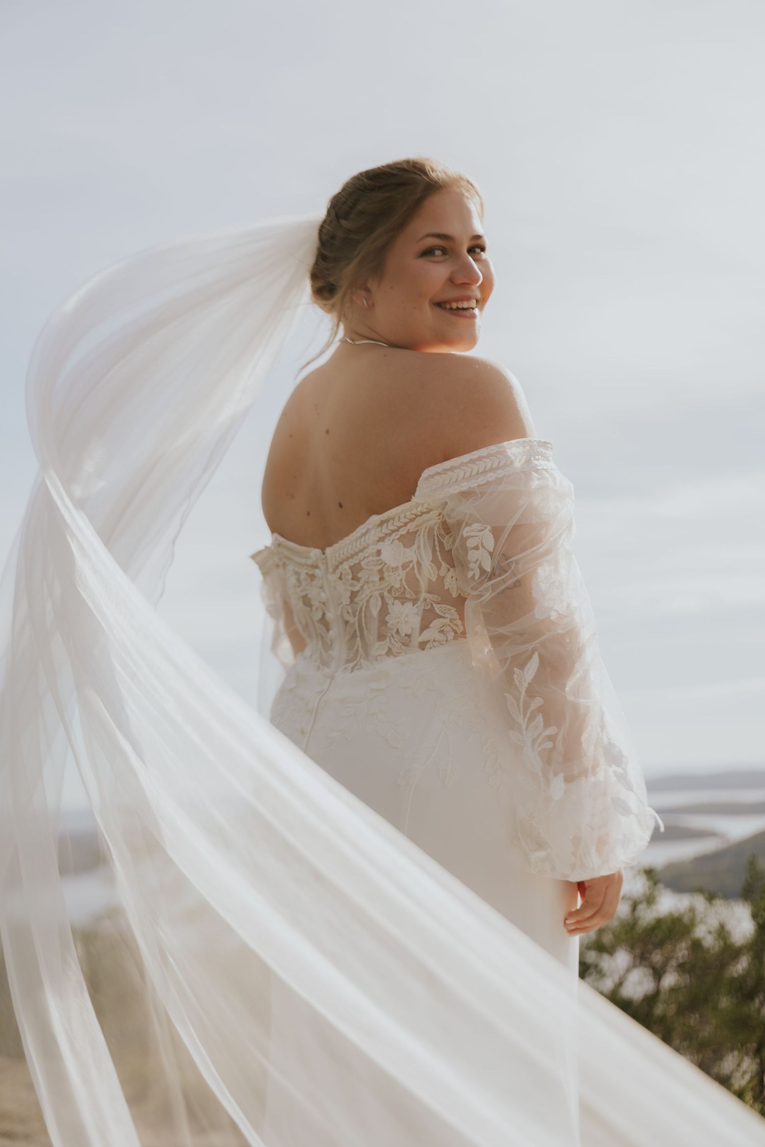 Bride in off-shoulder lace wedding dress with flowing veil poses against cloudy sky.