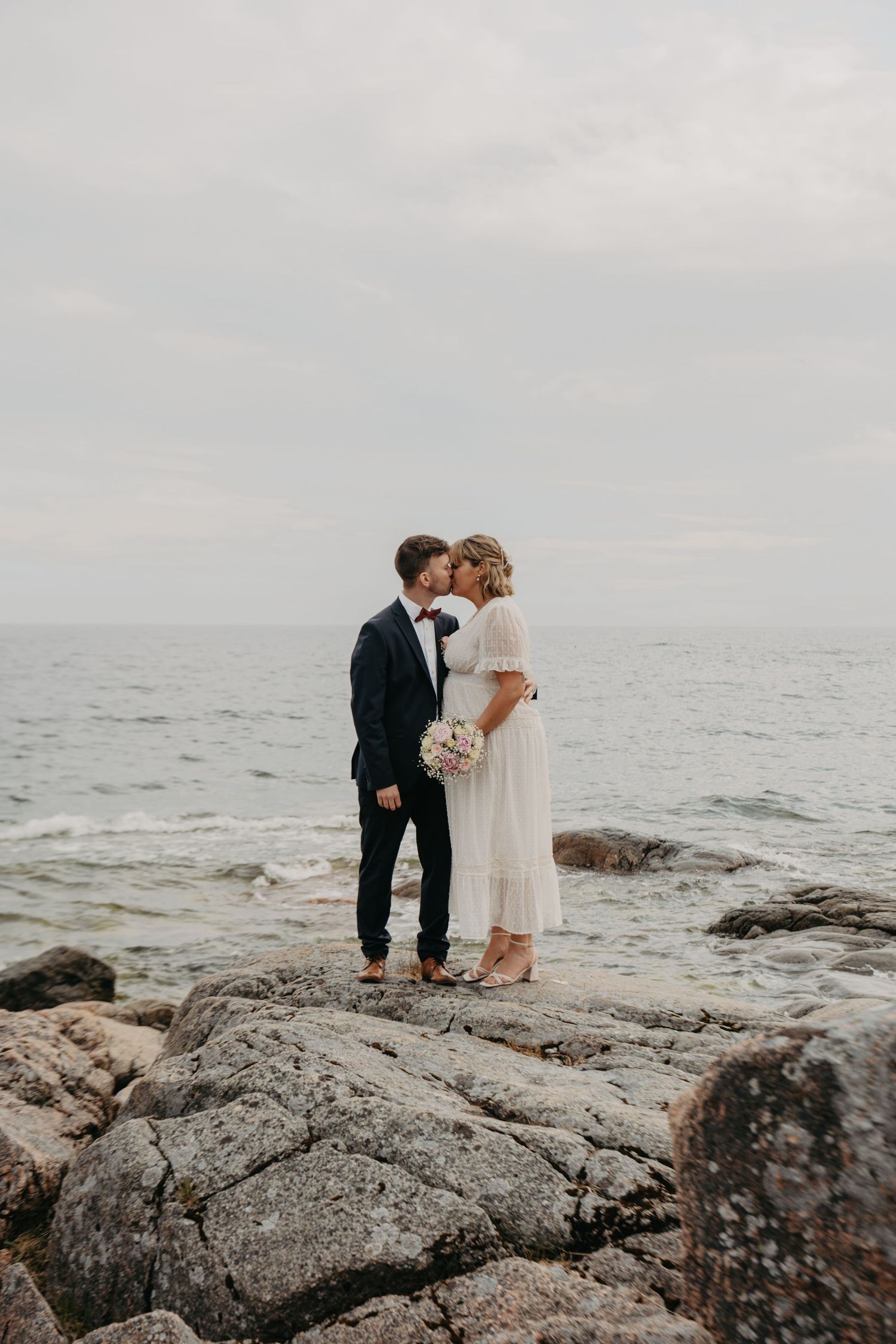 A couple in wedding attire embraces on dramatic rocky coastline under overcast skies.