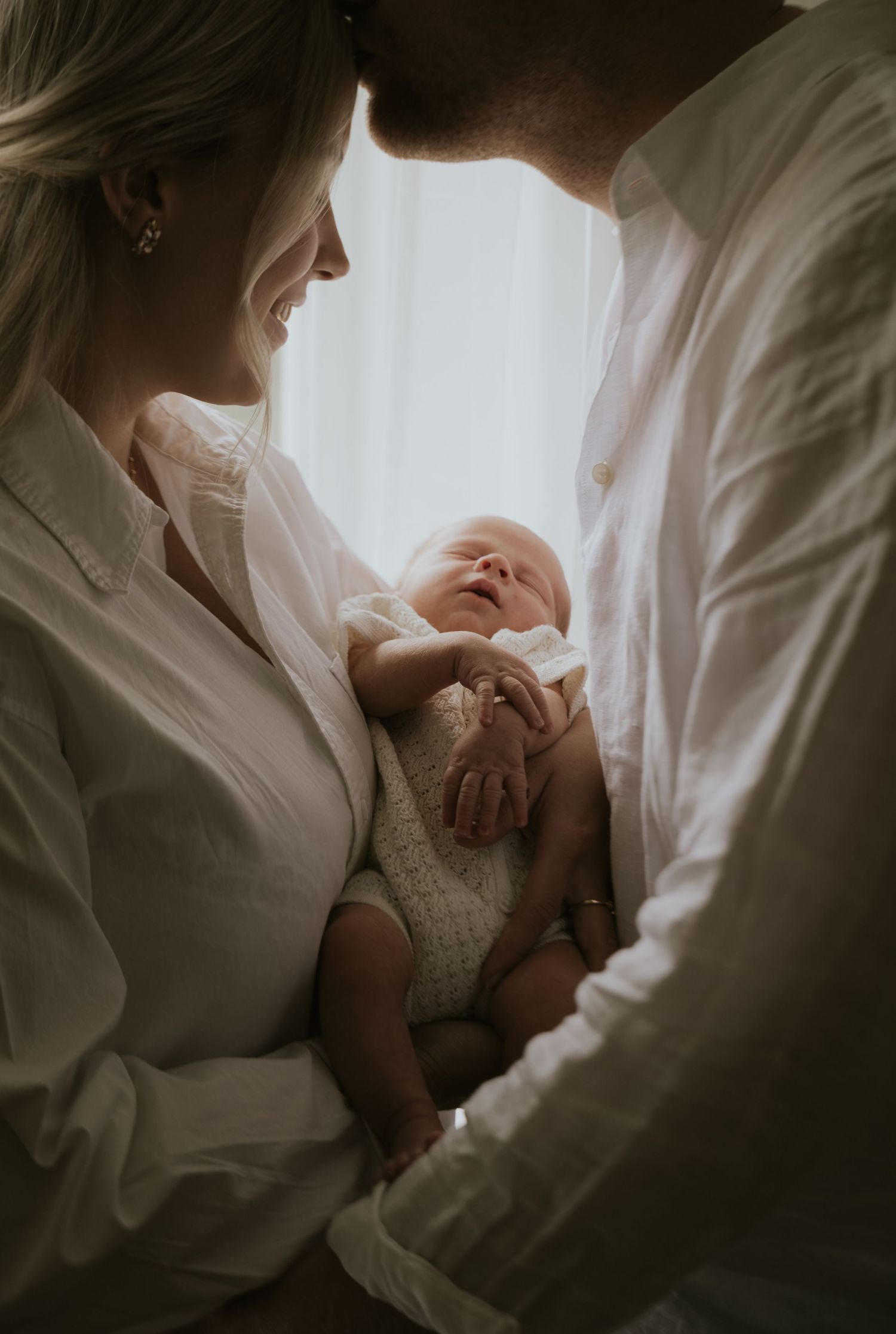 A tender moment between parents and their newborn baby in soft natural light against white curtains.