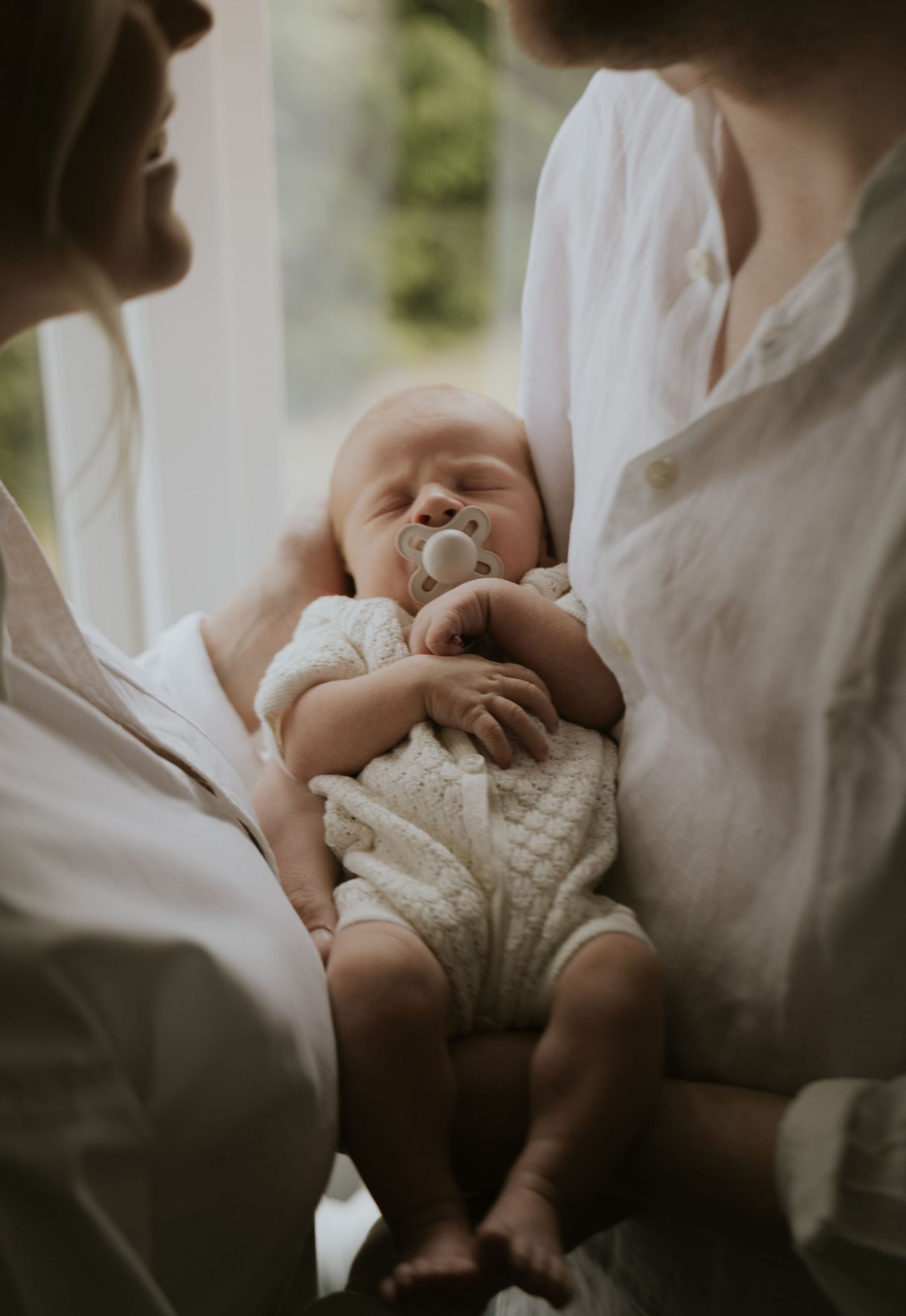 Soft natural light illuminates gentle moments between parents cradling their newborn in white clothing.