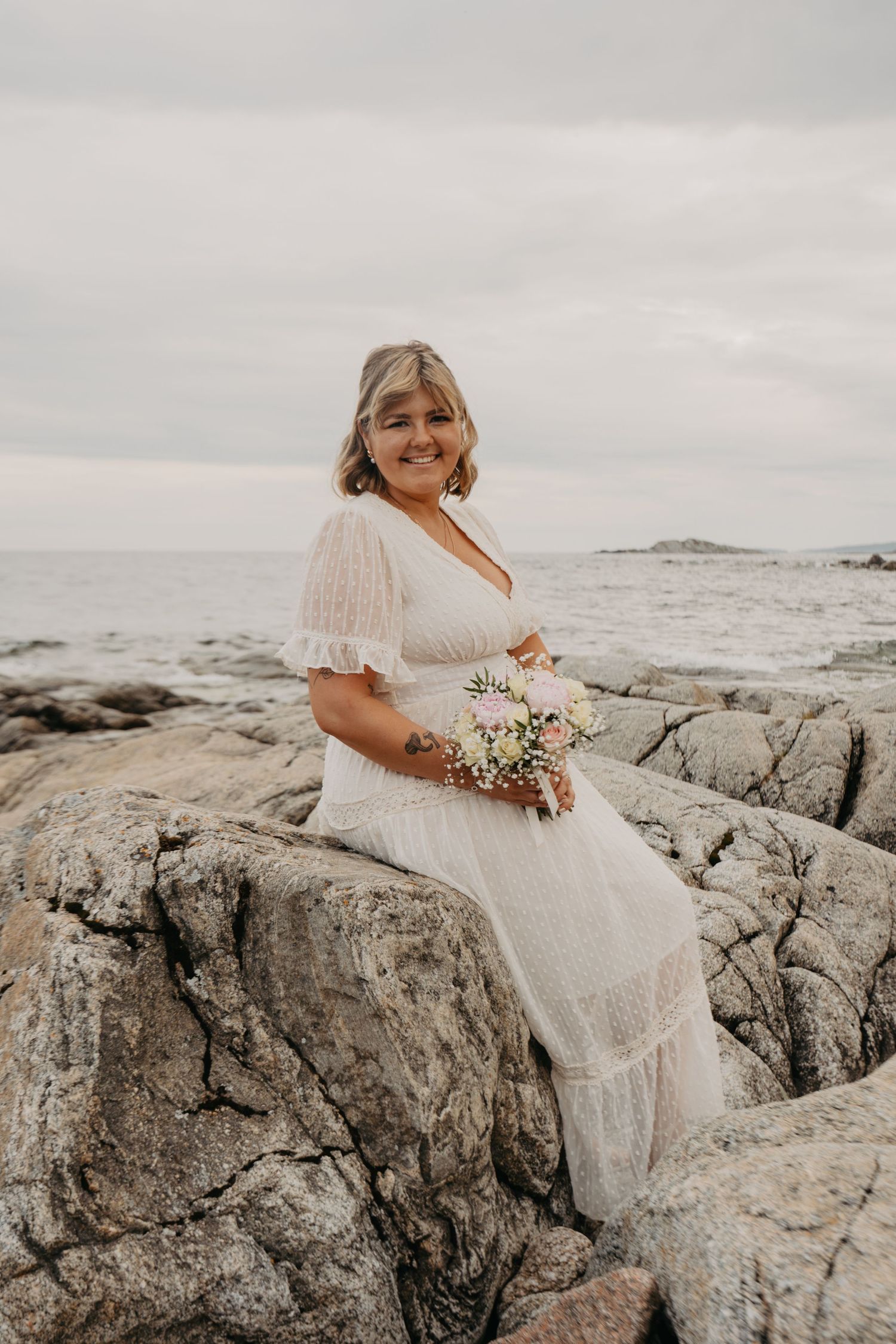 Bride in vintage-style lace dress poses on rocky coastline holding delicate floral bouquet.