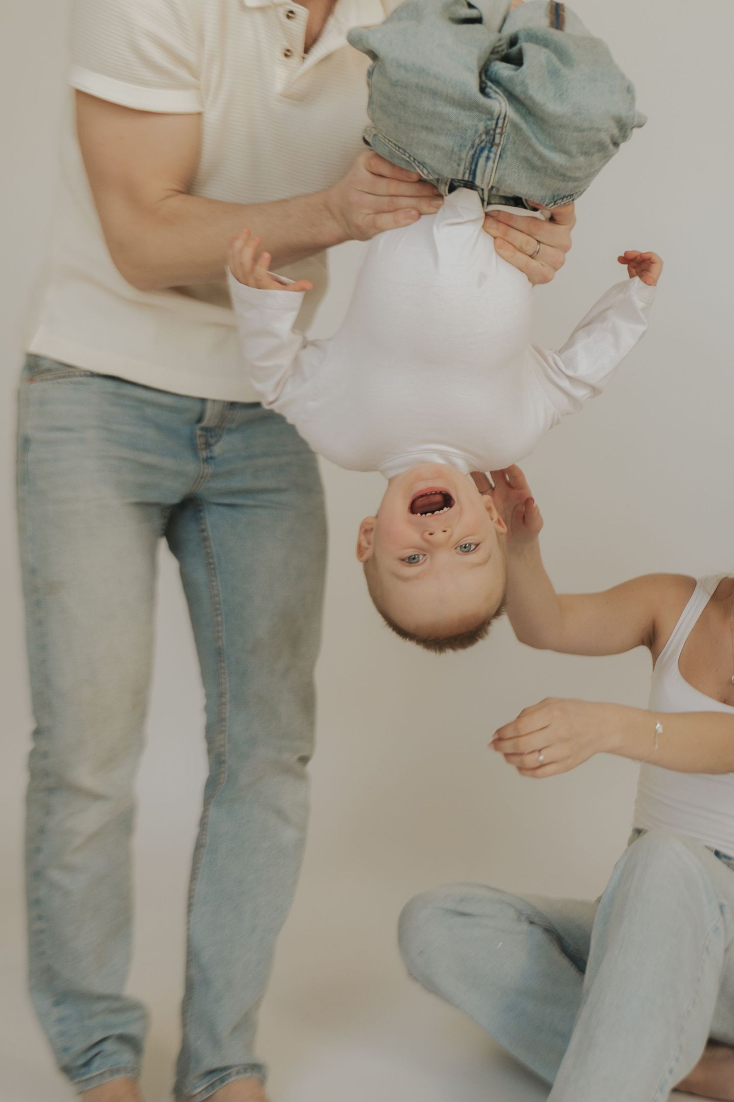 Playful family moment with baby being held upside down while wearing white onesie and jeans.