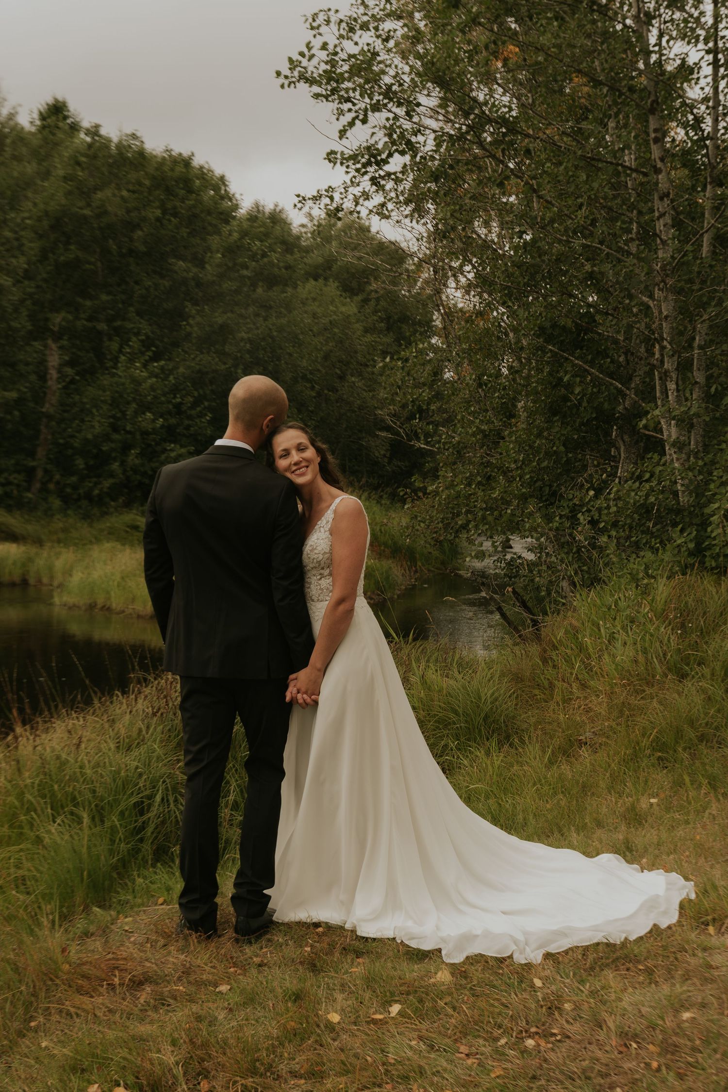 Wedding couple standing by lake with flowing white dress and forest backdrop.