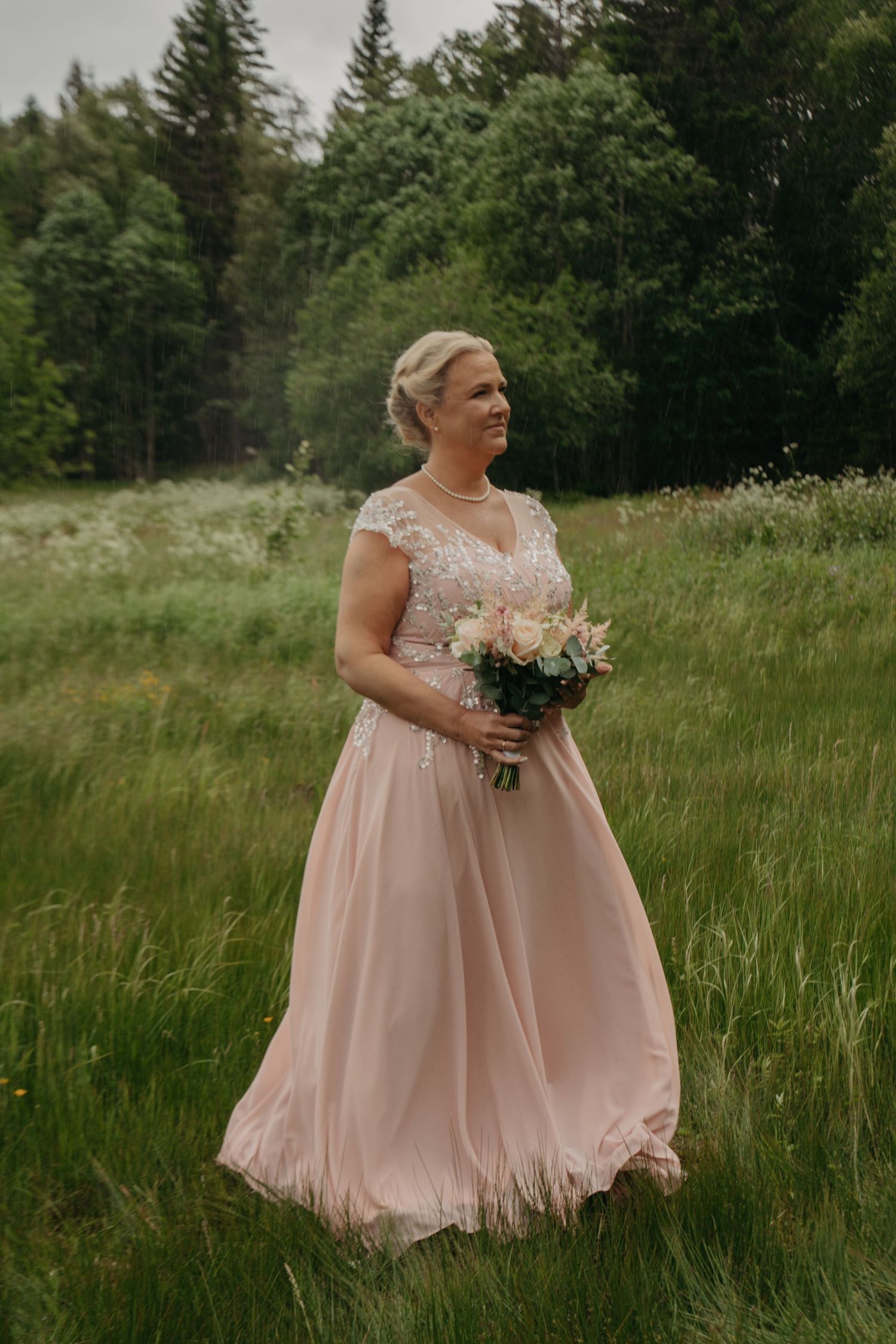 A bride in a blush pink lace gown holds a bouquet while standing in a grassy meadow.