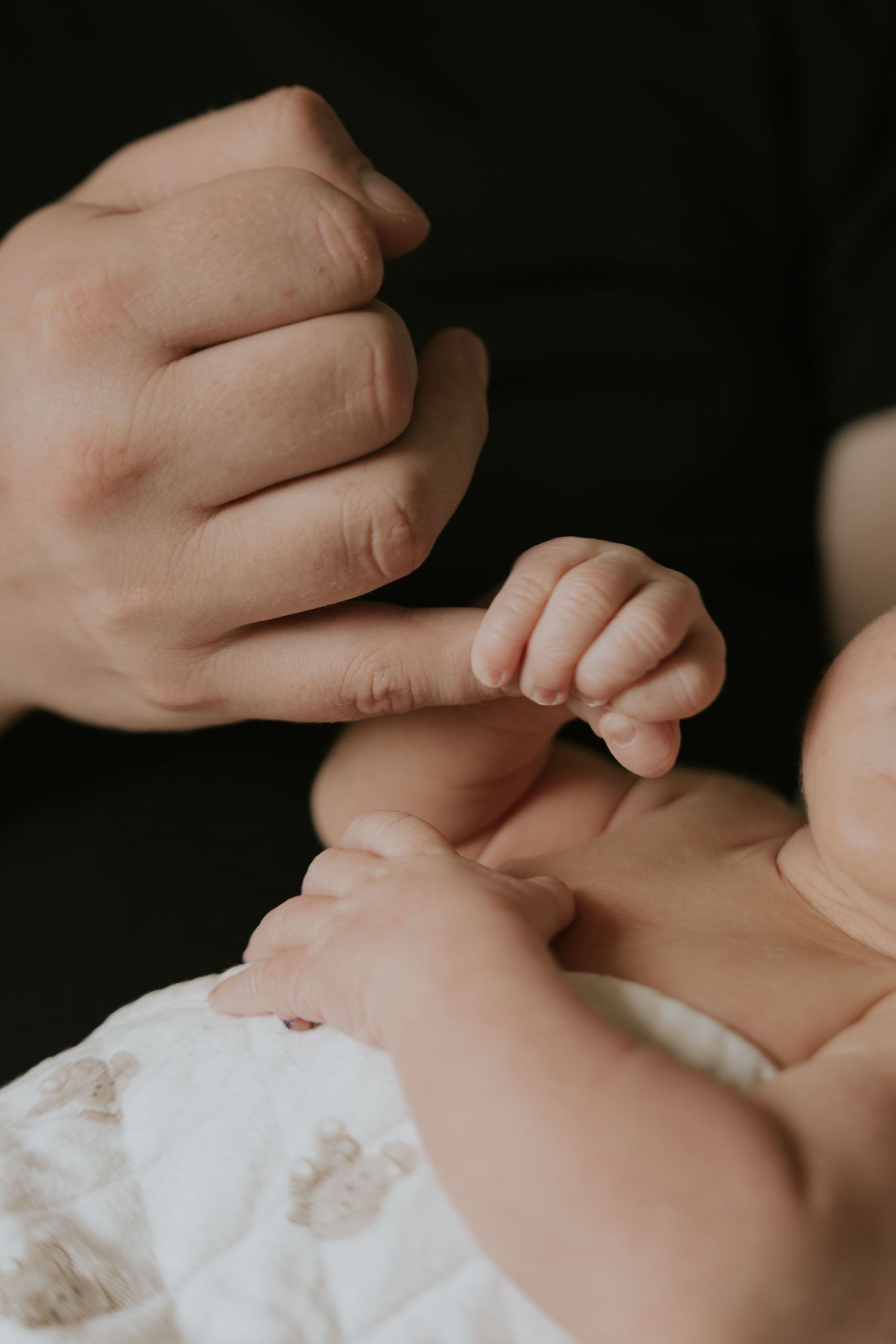 A close-up view of an adult's hand gently holding a baby's tiny hand against a dark background.