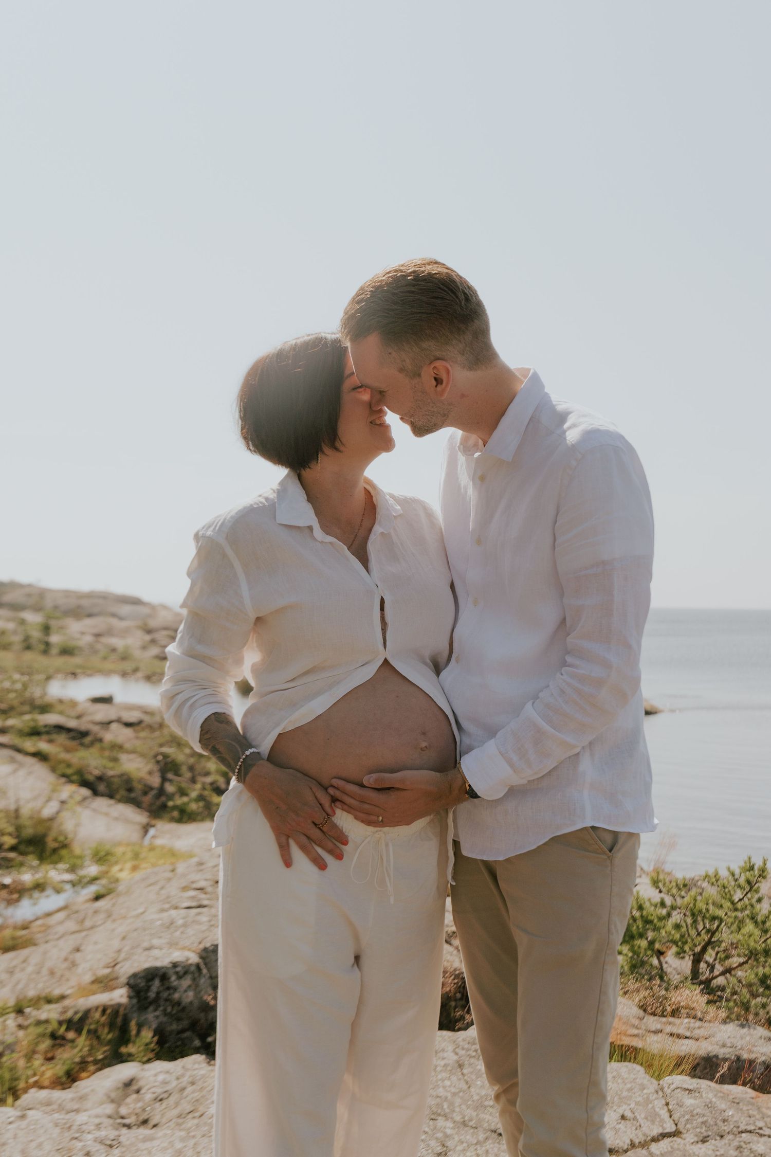 A couple dressed in white shares tender moments along a rocky coastline during a sunny day.