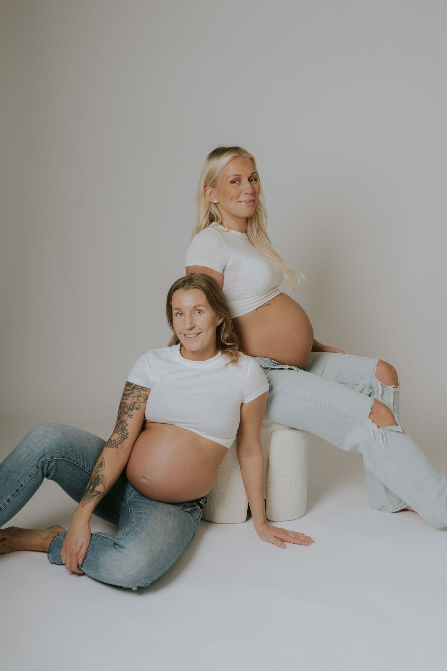 Two pregnant women in casual attire lounging on white background in artistic poses.