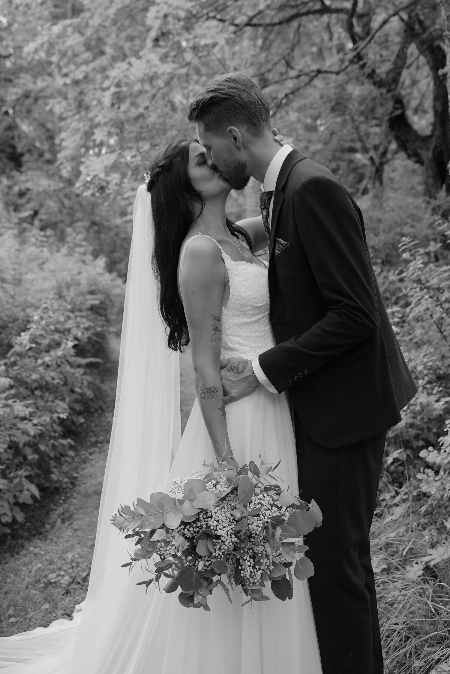 Black and white wedding portrait with couple holding bouquet in forest setting.