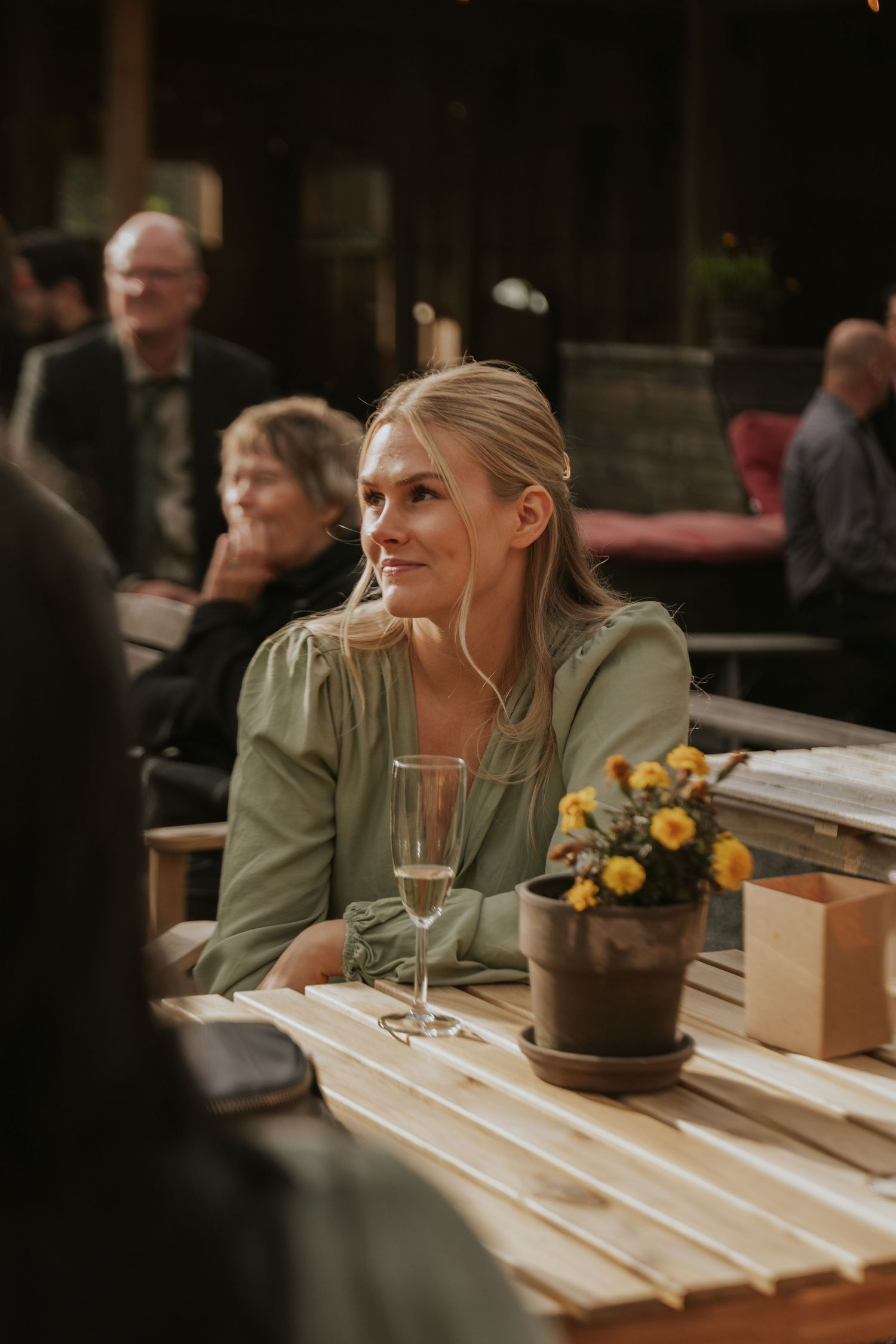 Person in olive green shirt sits at wooden restaurant table with wine glass and floral centerpiece.